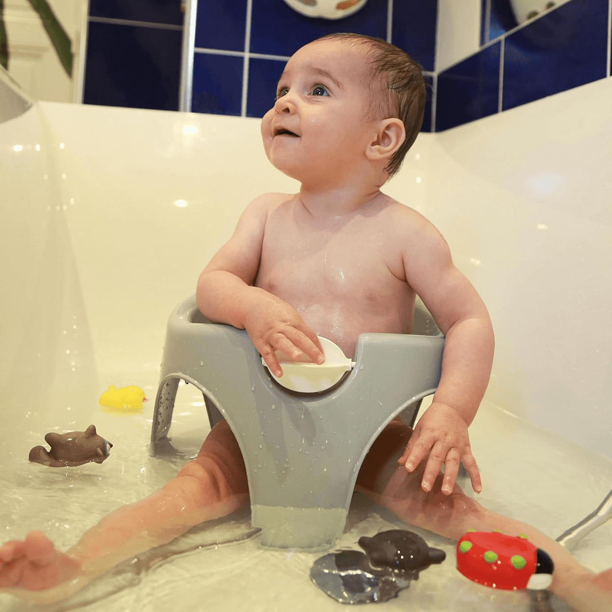 Un bebé feliz se sienta en un asiento de baño gris dentro de una tina blanca, rodeado de juguetes.