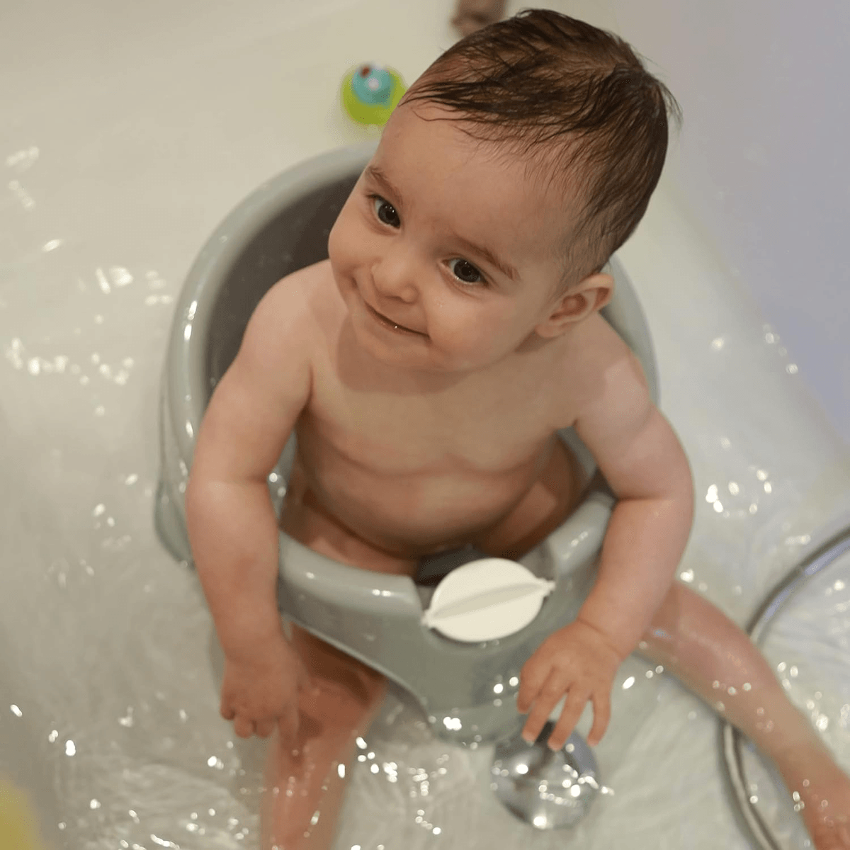 Un bebé feliz se sienta en un asiento de baño gris en una tina llena de agua, mirando hacia arriba.
