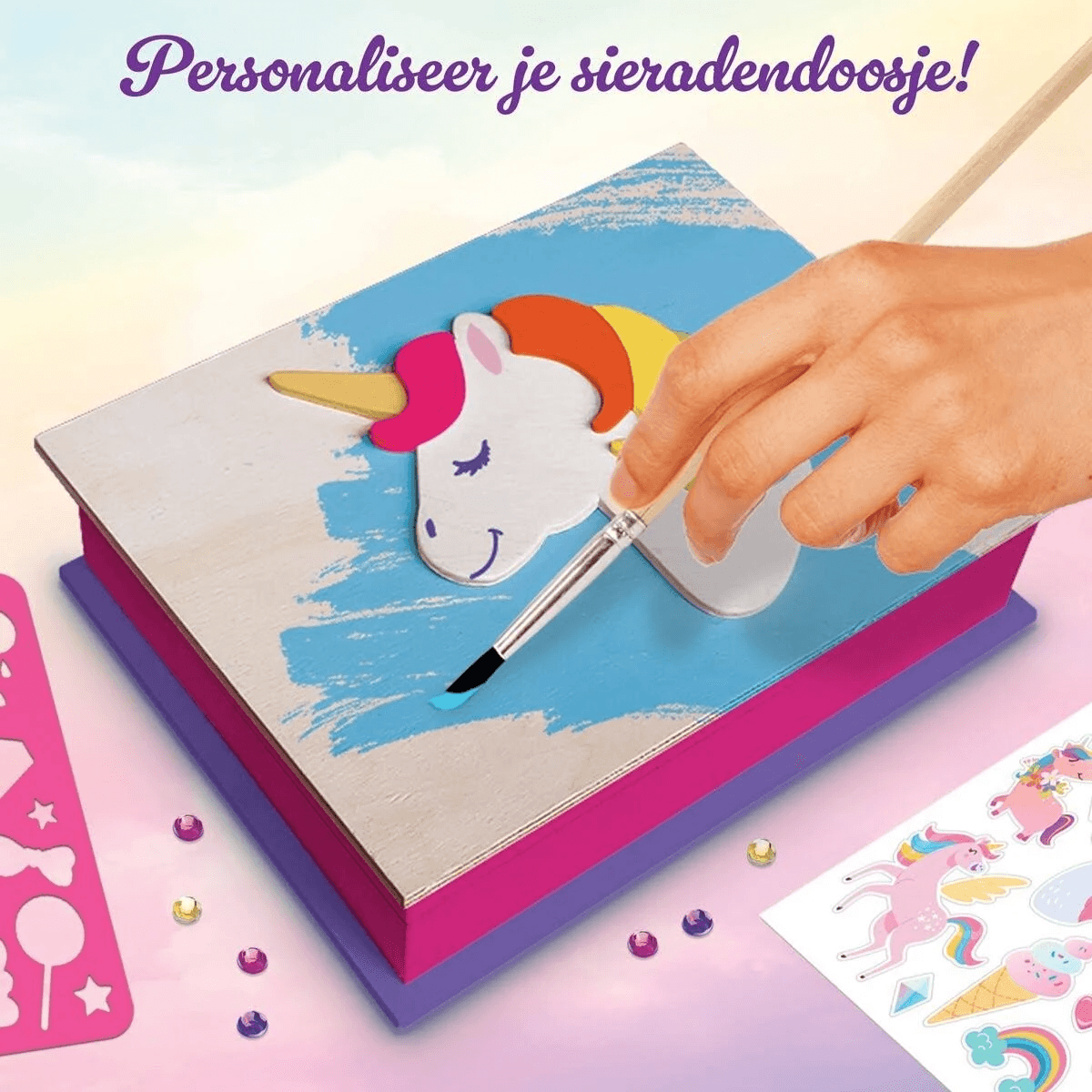 Una mano pinta una caja de unicornio con pintura azul, rodeada de pegatinas y gemas.