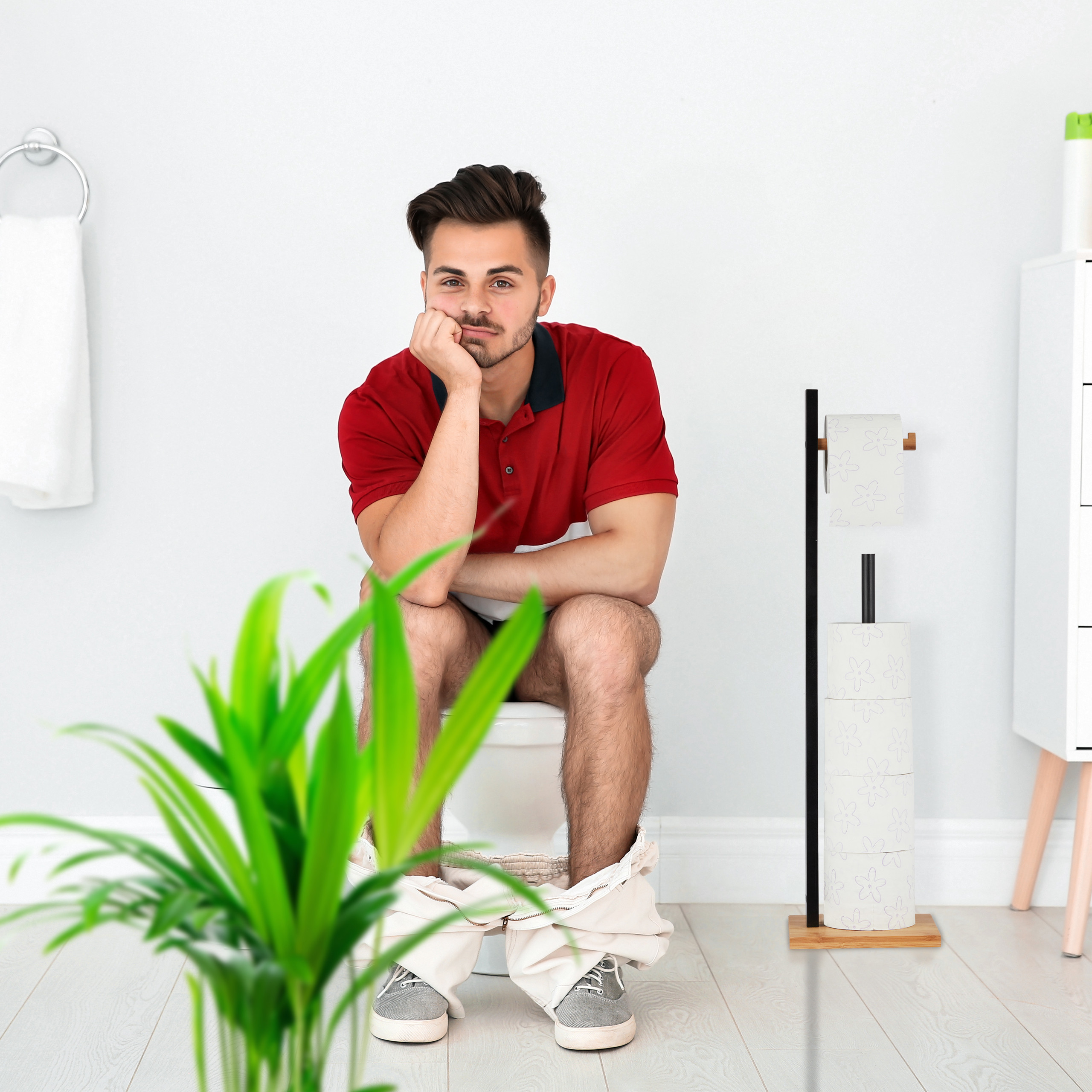 Ein Mann sitzt auf der Toilette, mit heruntergelassener Hose und st ützt den Kopf auf die Hand in einem hellen Badezimmer.
