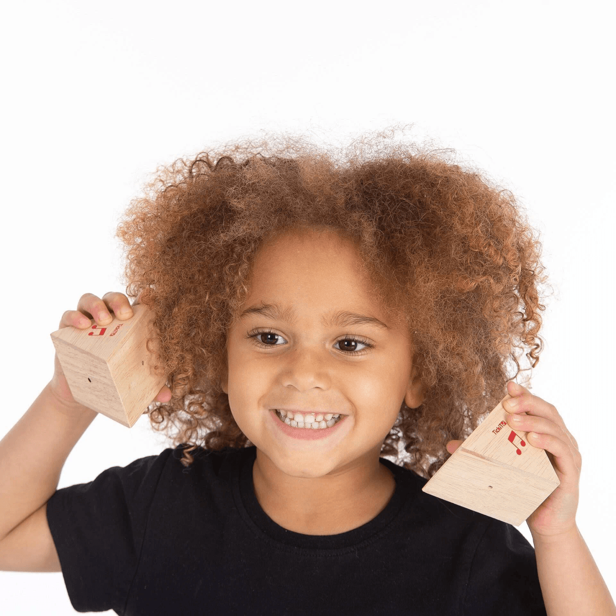 Un niño sonriente con cabello rizado sostiene bloques de madera cerca de sus orejas.