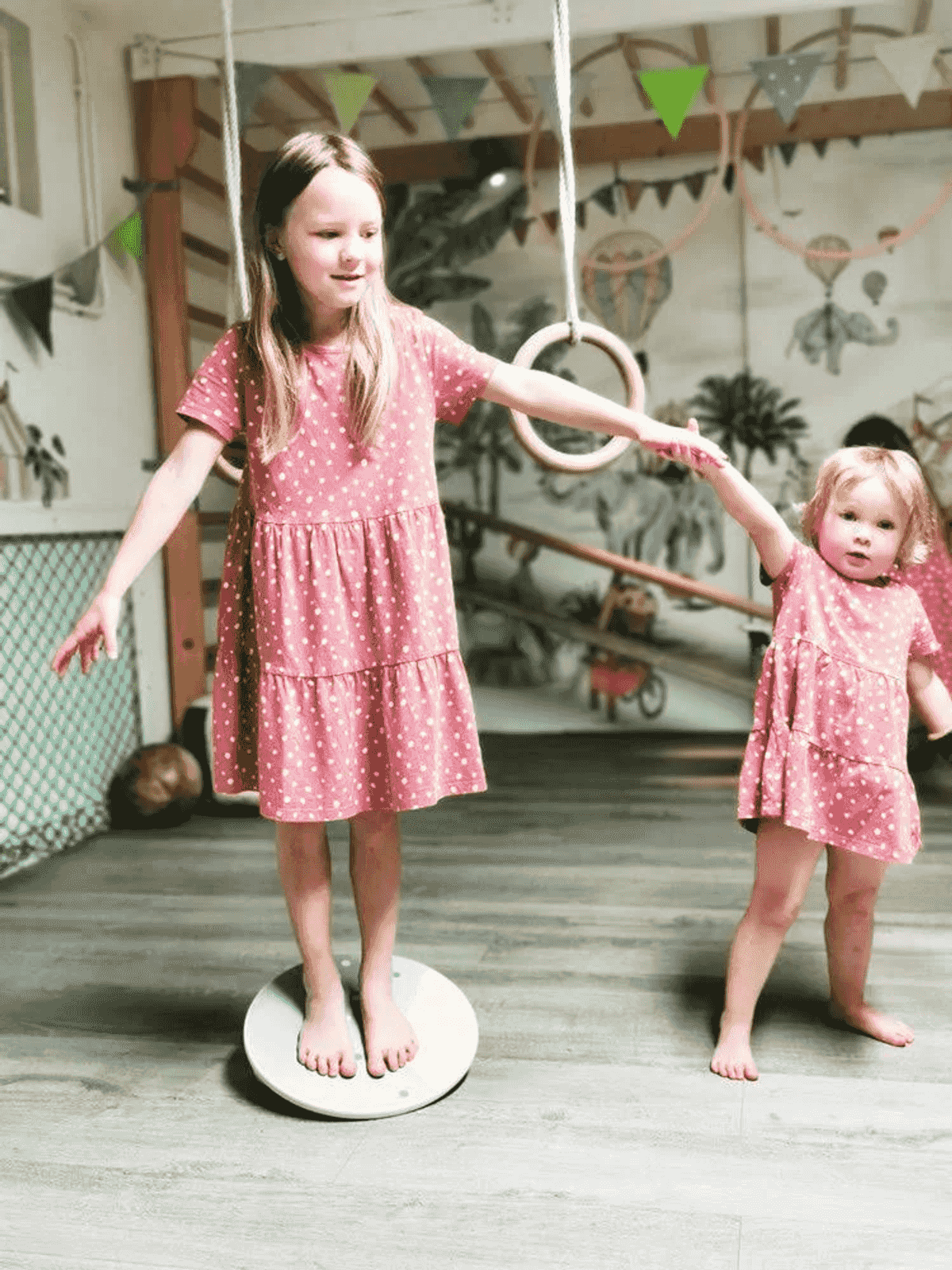 Dos niñas con vestidos rosados juegan en una sala de juegos, una equilibrándose en un disco redondo.