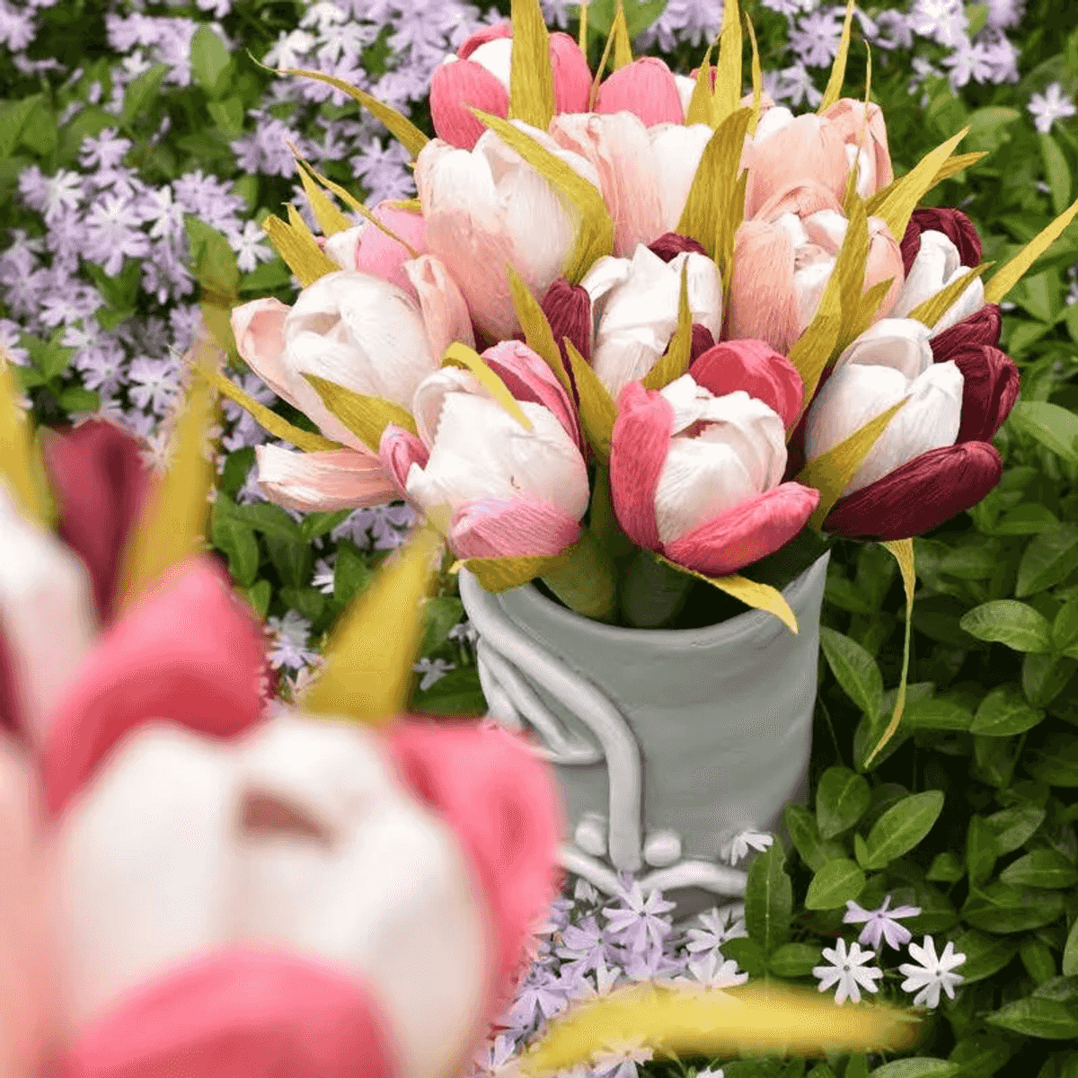 Una cesta de tulipanes de papel rosas y blancos al aire libre con pequeñas flores moradas.