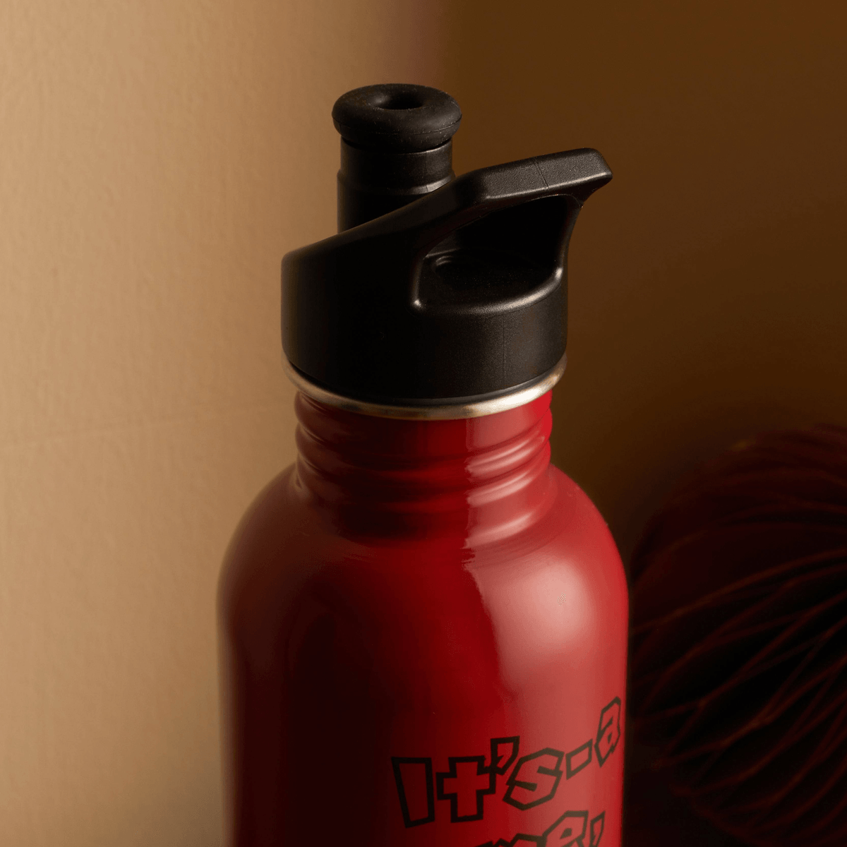 Una botella de agua de metal roja con tapa y asa negras, parcialmente visible contra un fondo cálido.