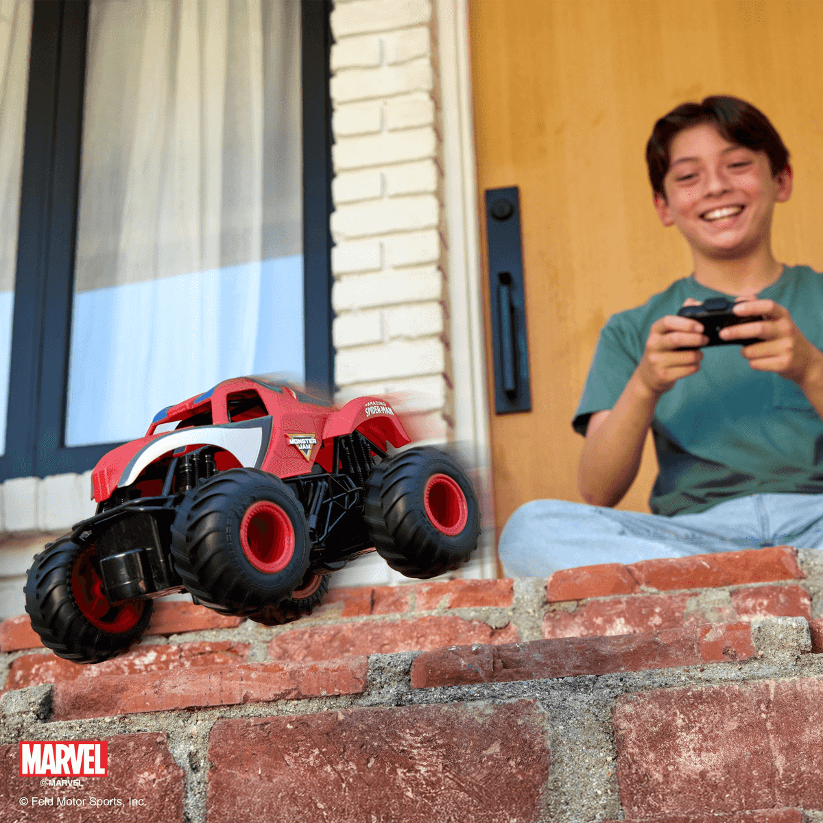 Un niño sonriente está sentado en unas escaleras y usa un control remoto para manejar un camión monstruo rojo.