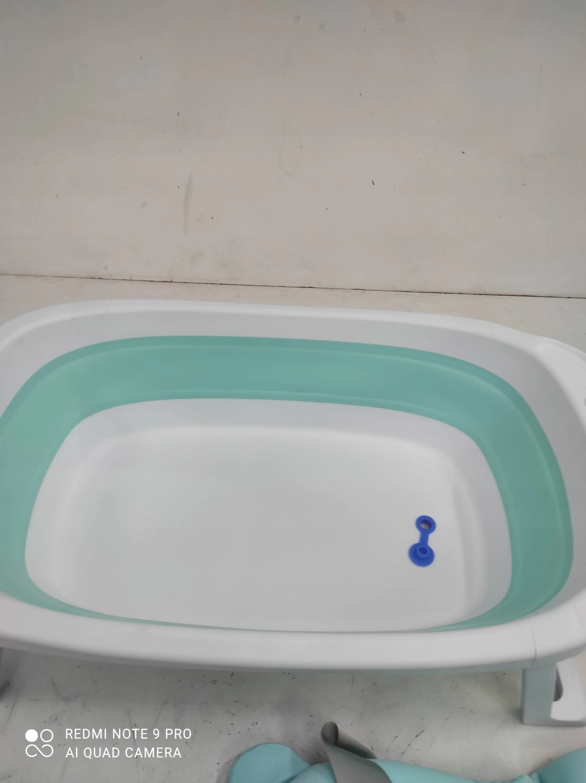 Una bañera plegable para bebé blanca y verde menta con un tapón azul sobre una superficie clara.
