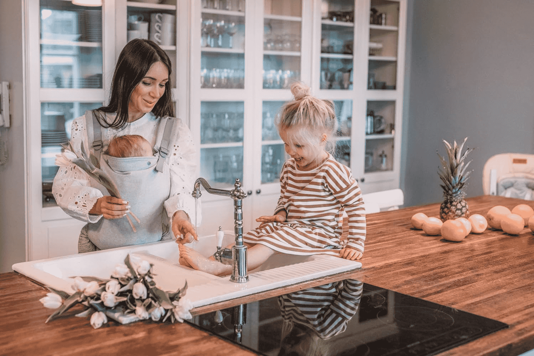 Una madre con un portabebés lava los pies de su hija en el fregadero de la cocina.