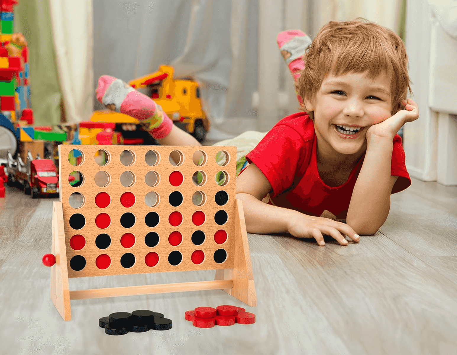 Un niño sonriente está acostado en el suelo jugando con un juego de cuatro en raya de madera.