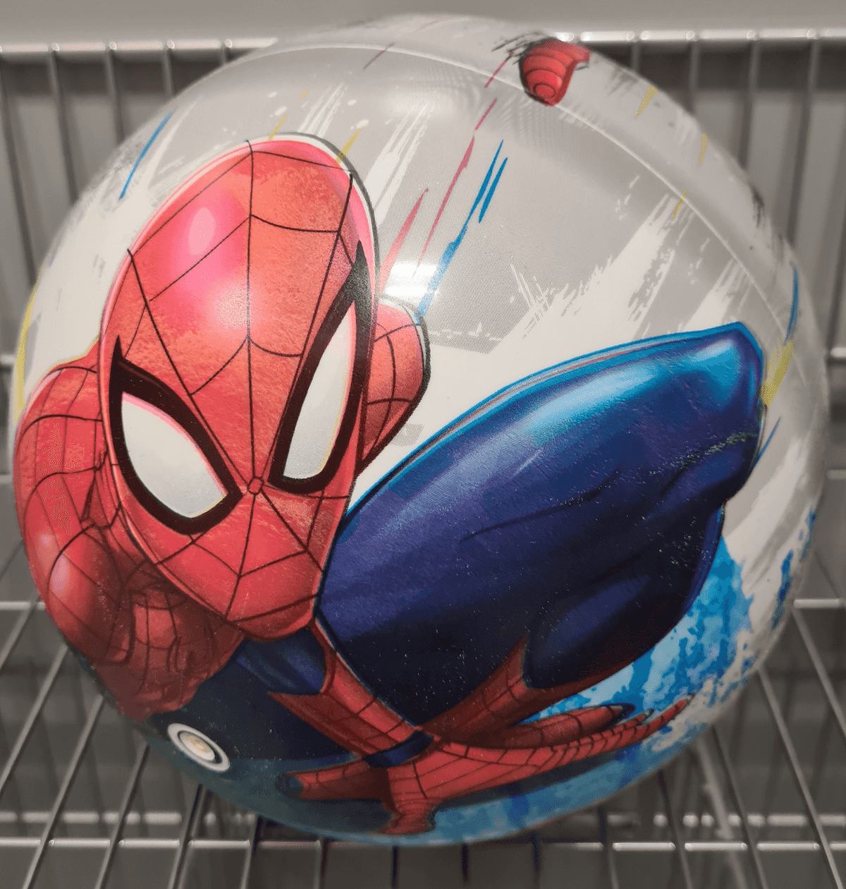 Una pelota de juguete redonda con una ilustración de Spider-Man en estilo cómic en rojo, azul y blanco sobre una rejilla metálica.