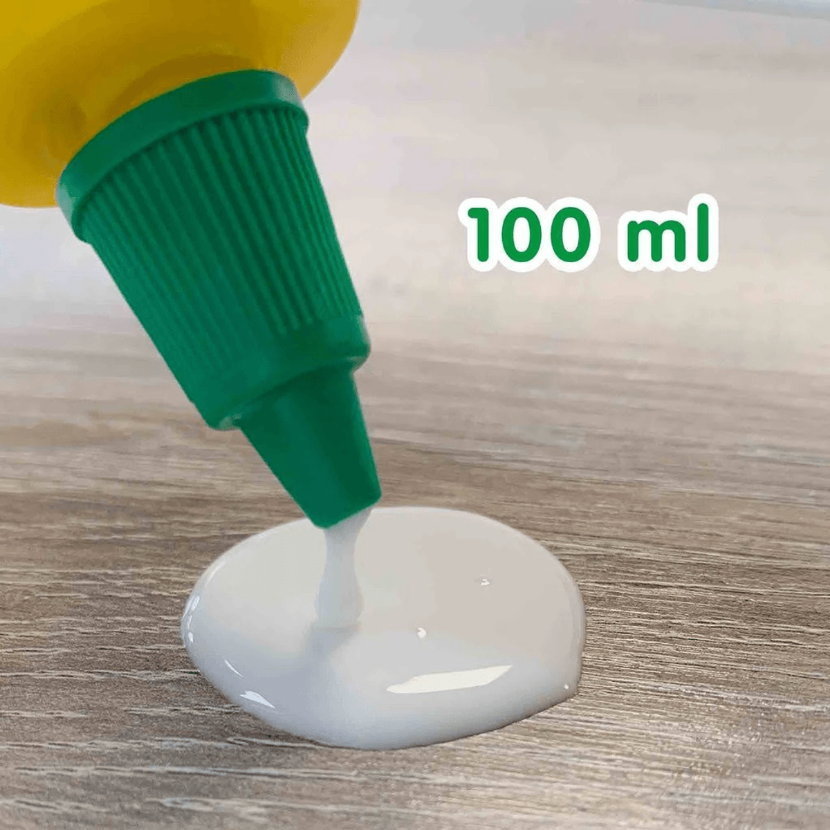 Un frasco de pegamento con tapa verde vierte pegamento blanco sobre una superficie de madera, con el texto 100 ml.