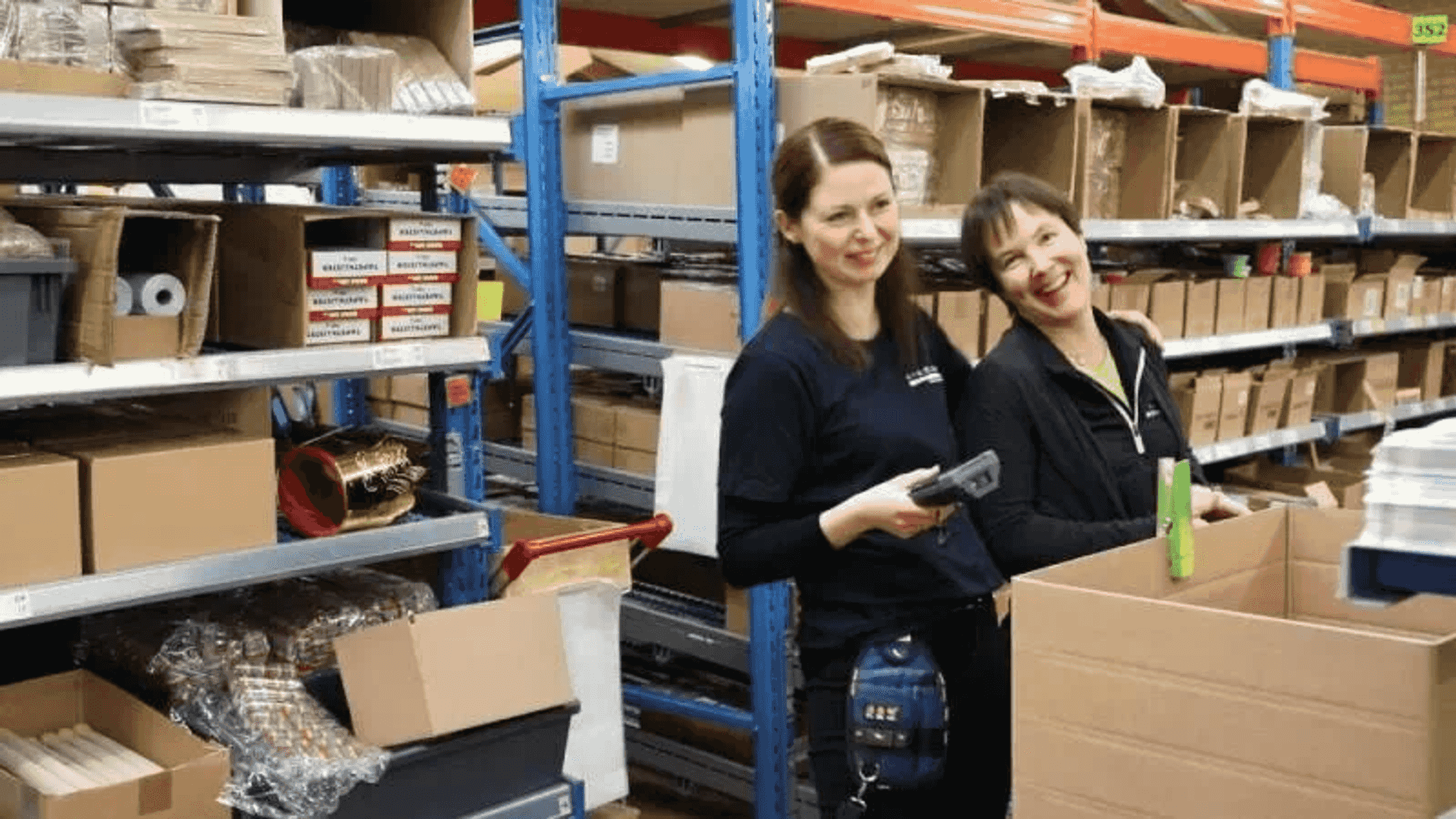 Dos empleadas sonrientes en un gran almacén con estantes llenos de cajas de cartón, escaneando artículos.