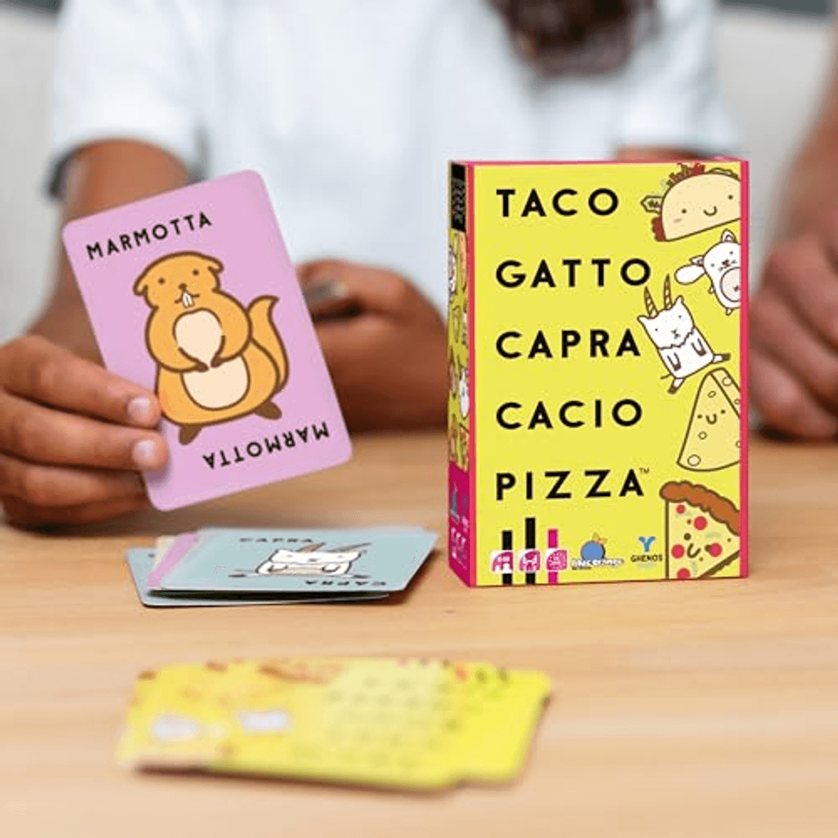 Una persona tiene in mano una carta rosa con una marmotta, accanto a una scatola di gioco gialla.