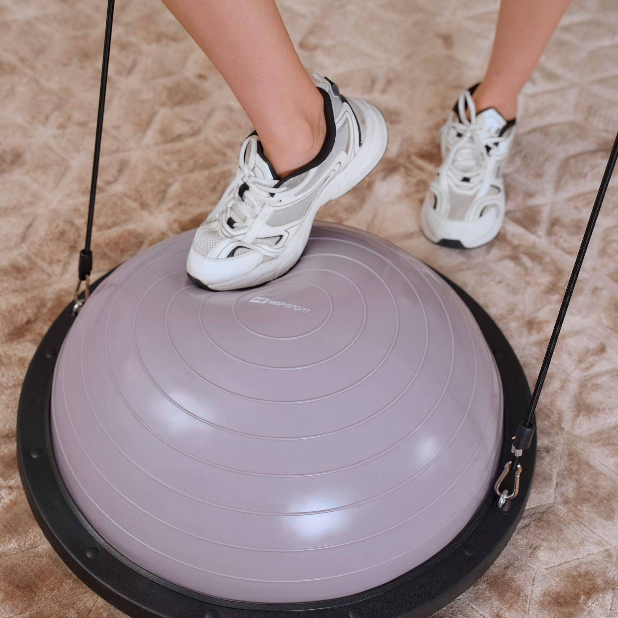 Eine Person mit weißen Turnschuhen steht auf einem violetten Balance Ball auf einem Teppichboden.