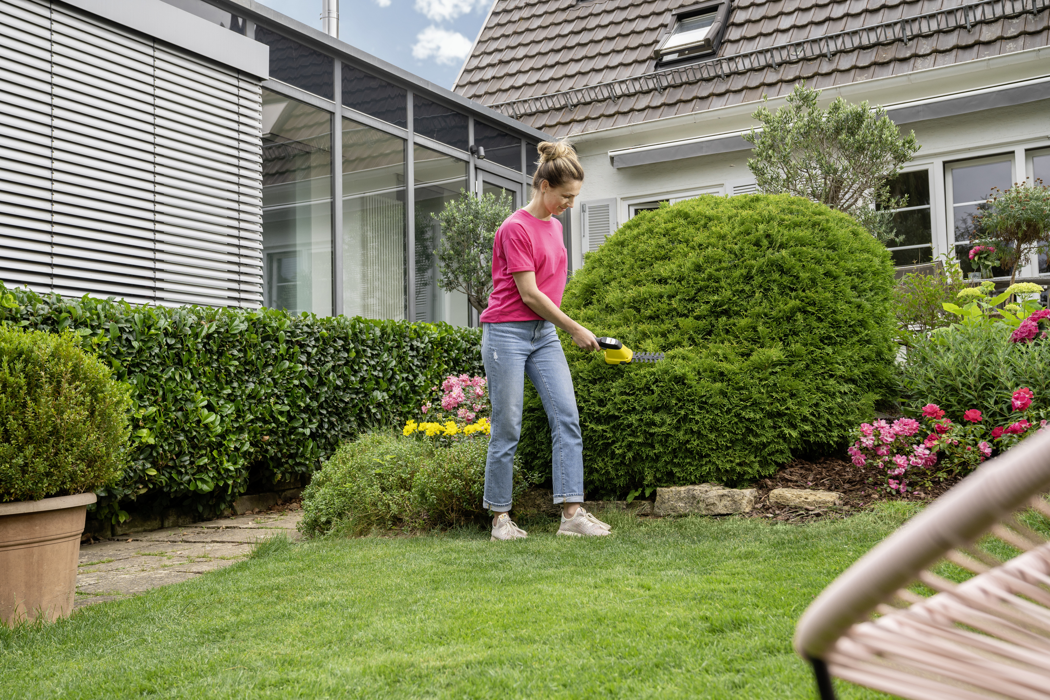 Eine Frau in pinkem Shirt und Jeans schneidet eine große Hecke mit einer elektrischen Heckenschere im Garten.