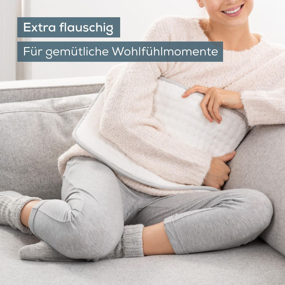 Eine Frau in gemütlichem Pullover und grauer Hose sitzt auf einem Sofa und drückt ein weißes Heizkissen an.
