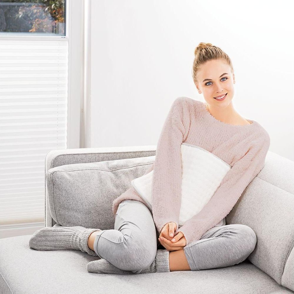 Eine lächelnde Frau in rosa Pullover und grauer Hose sitzt auf einem Sofa und hält ein weißes Heizkissen.