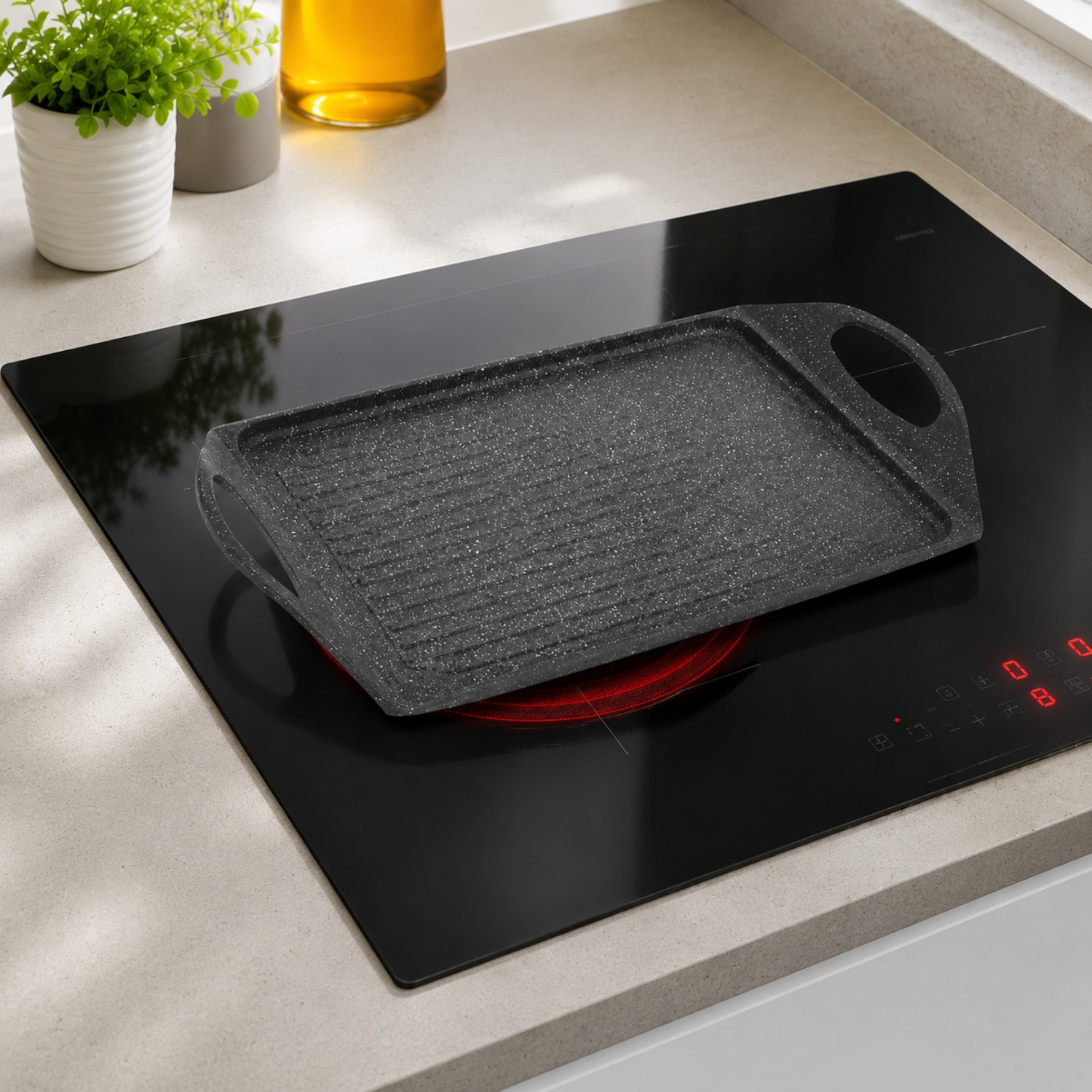 Una bistecchiera grigia con manici si trova su un piano cottura a induction nero in una cucina luminosa.