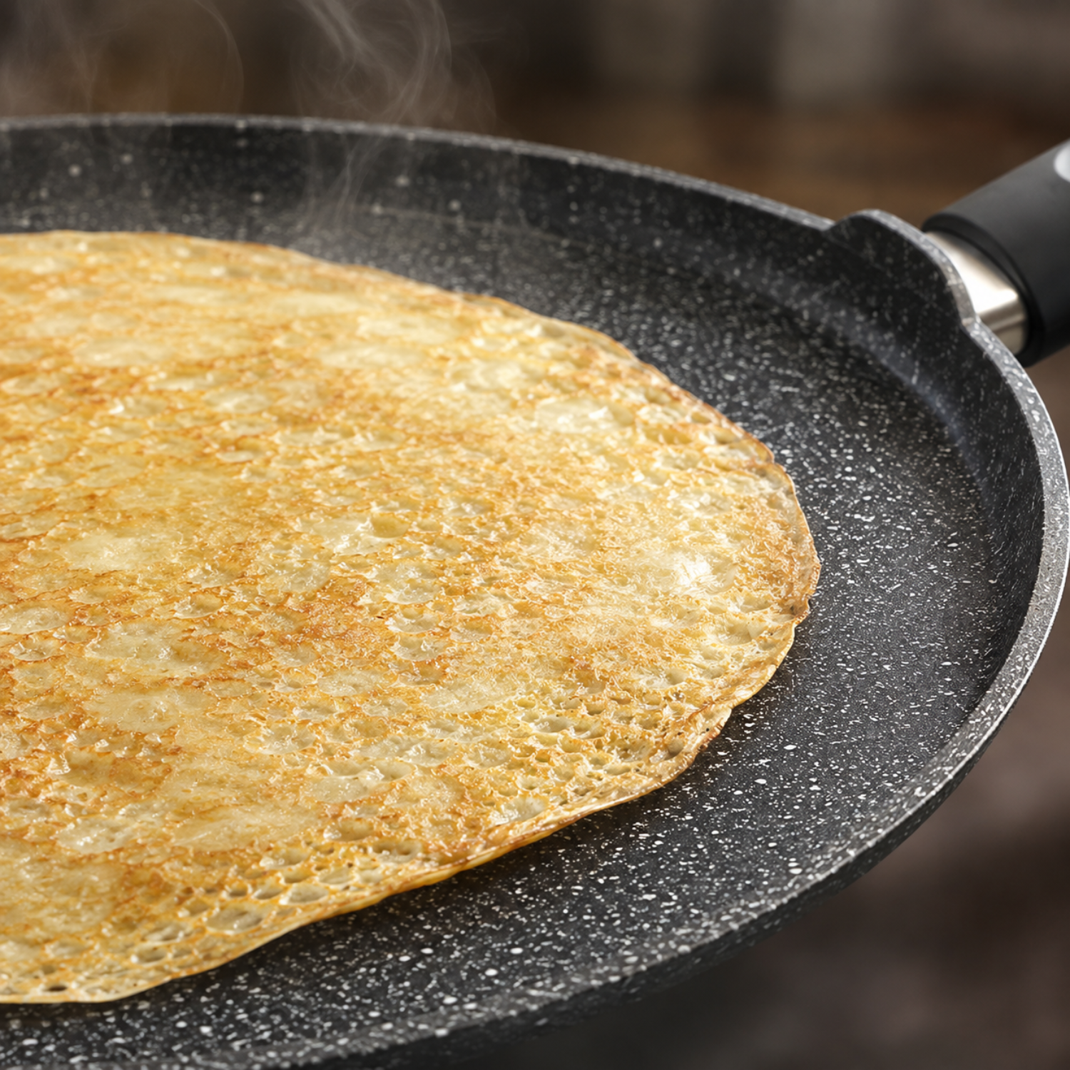 Una crepe dorata e fumante si cuoce in una padella nera marezzata.