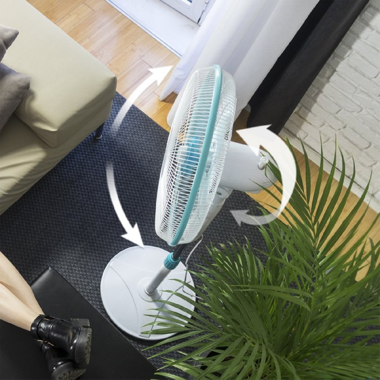 Un ventilador de pie oscilante blanco se encuentra en una sala de estar, cerca de un sofá, una planta y una ventana.