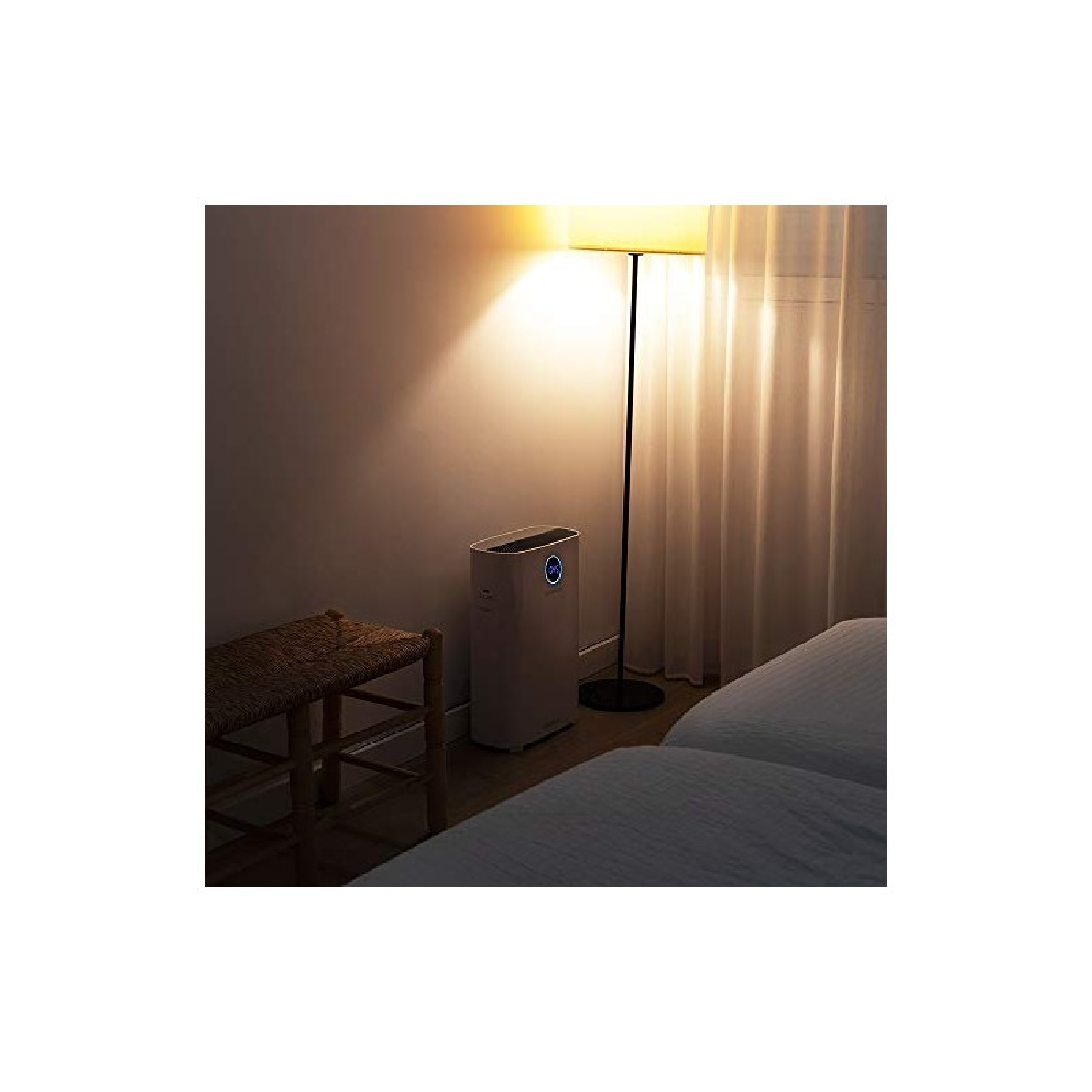 Un purificatore d'aria bianco si trova in una camera da letto accanto a un letto, uno sgabello e una lampada.