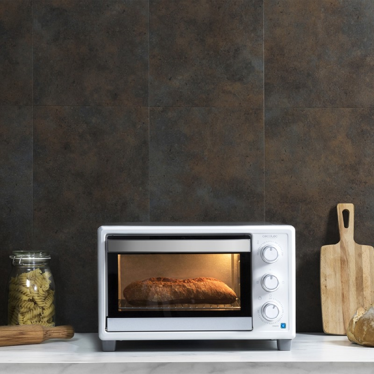 Un mini forno bianco su un ripiano cuoce il pane, con pasta, un tagliere e dell'impasto vicini.