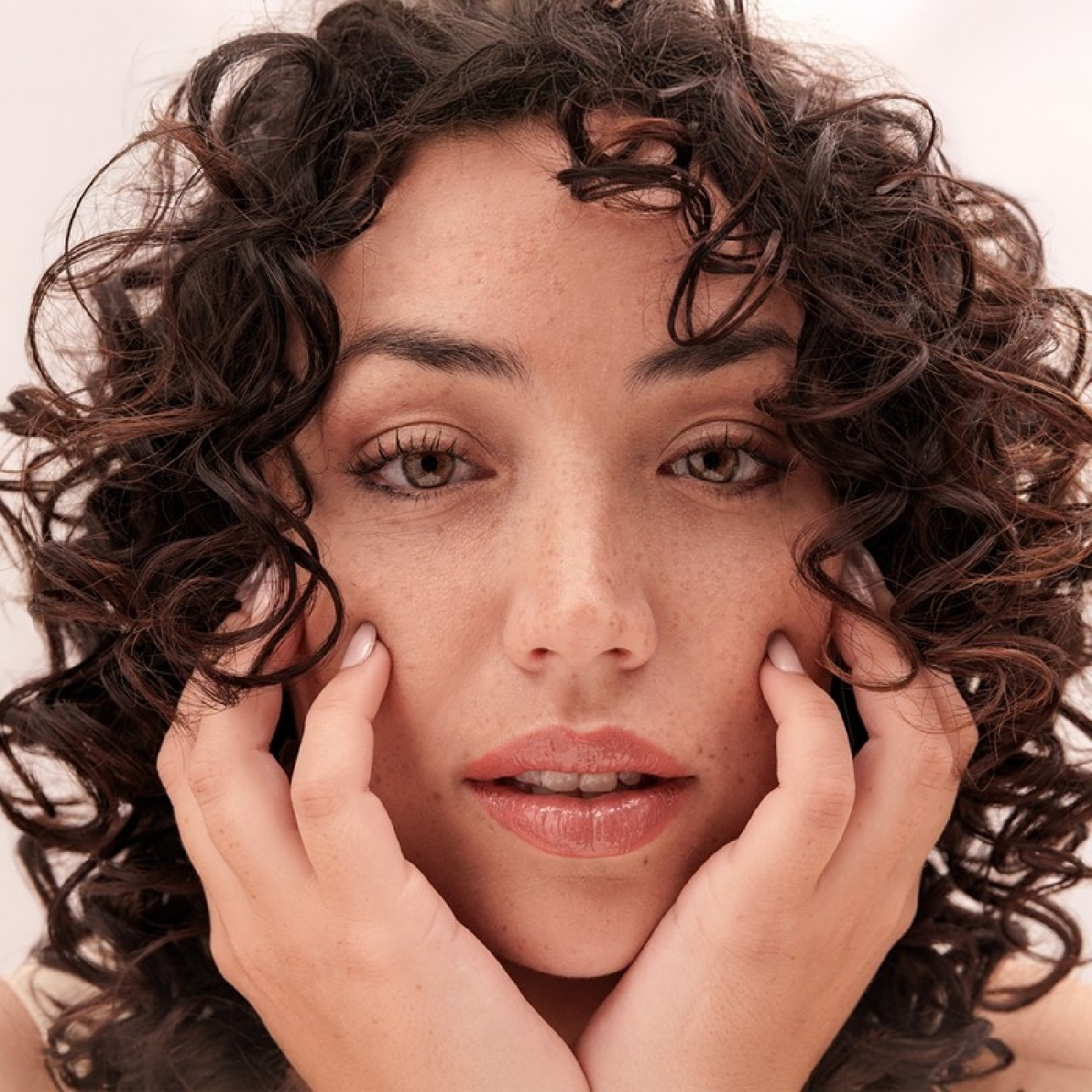 Primo piano di una donna con capelli ricci scuri e lentiggini che si tiene il viso con le mani.