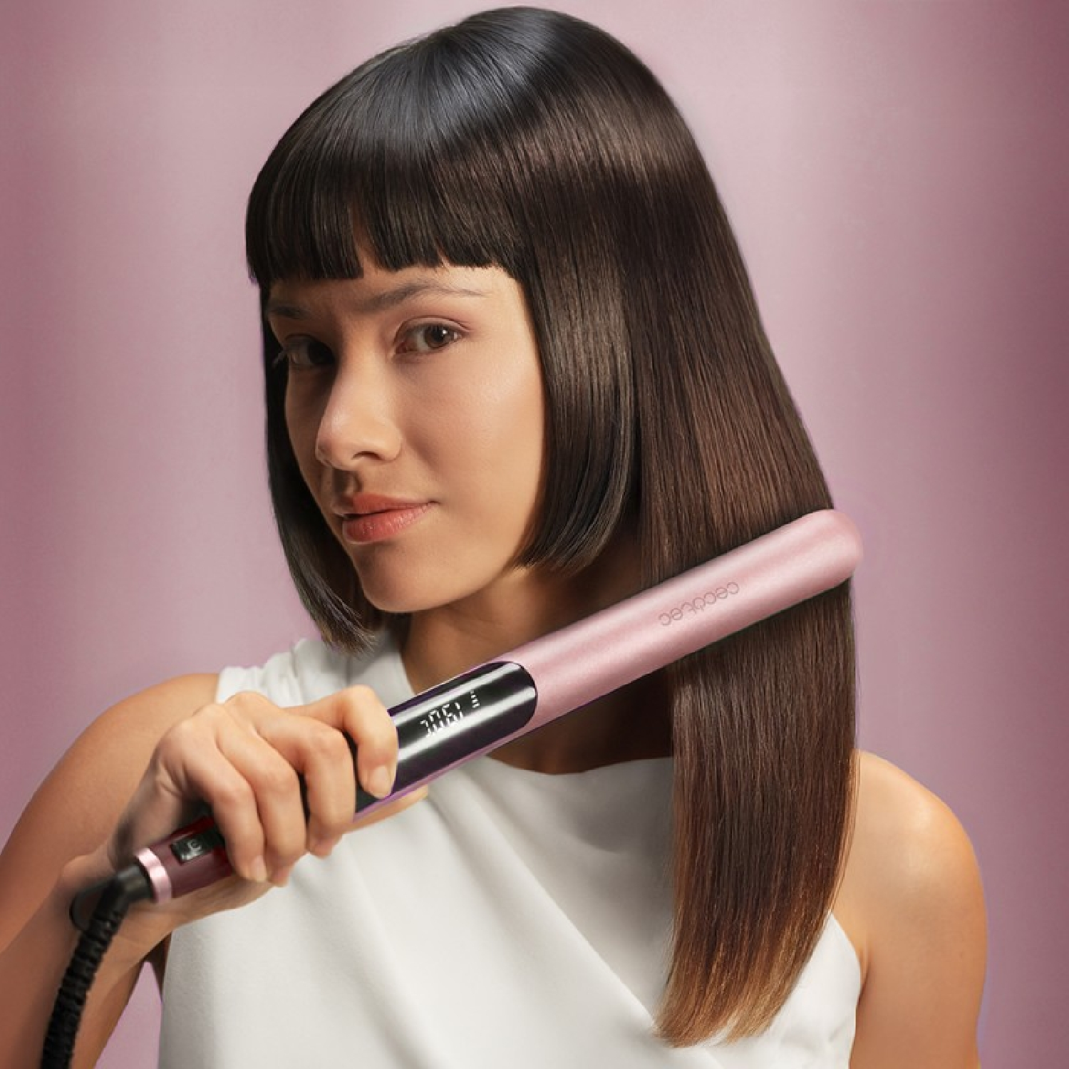 Donna con capelli castani lisci tiene una piastra per capelli rosa su sfondo rosa.