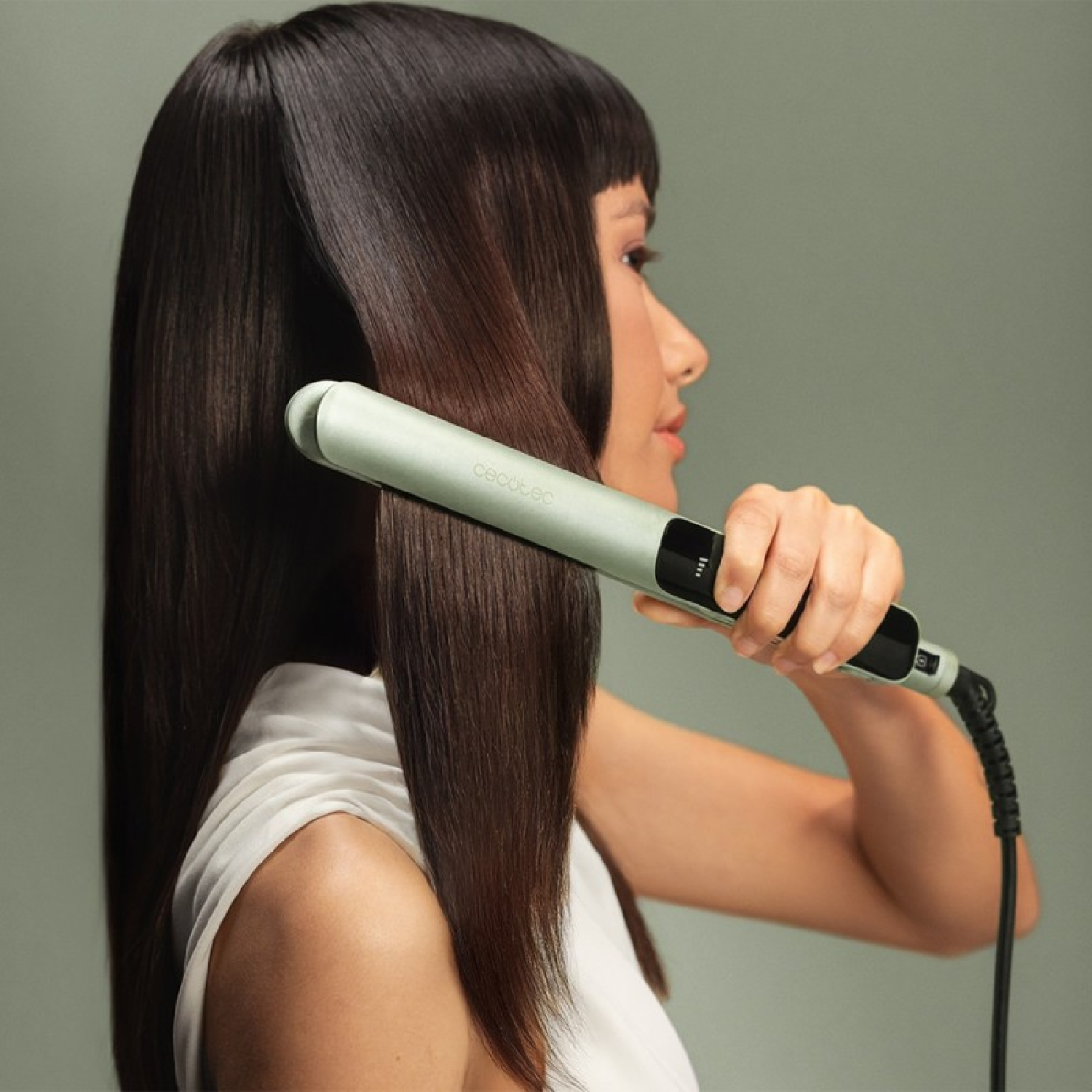 Una donna liscia i suoi lunghi capelli scuri con una piastra verde chiaro su sfondo grigio.