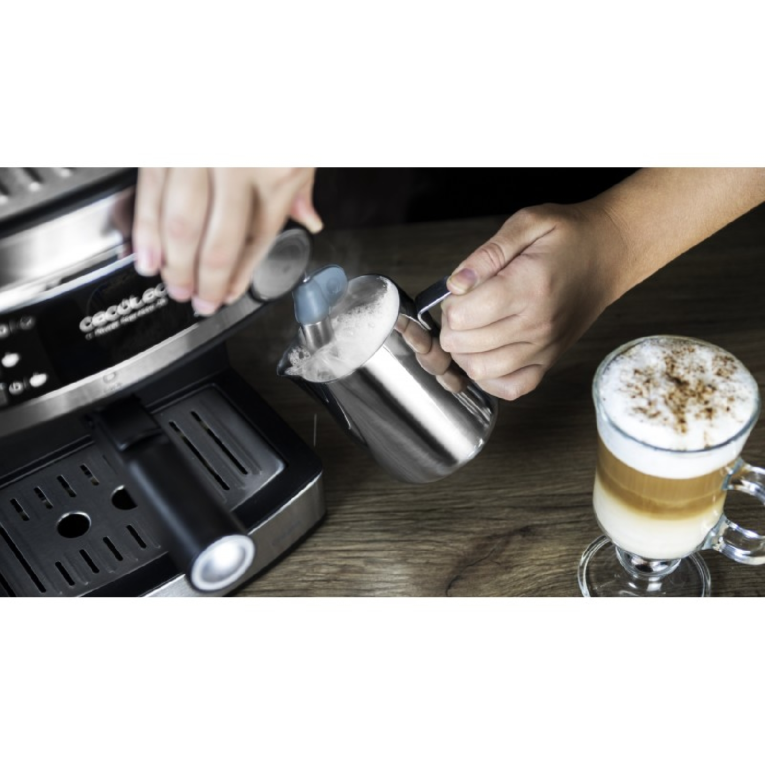 Alguien espuma leche en una jarra junto a una máquina de café y una taza de capuchino.