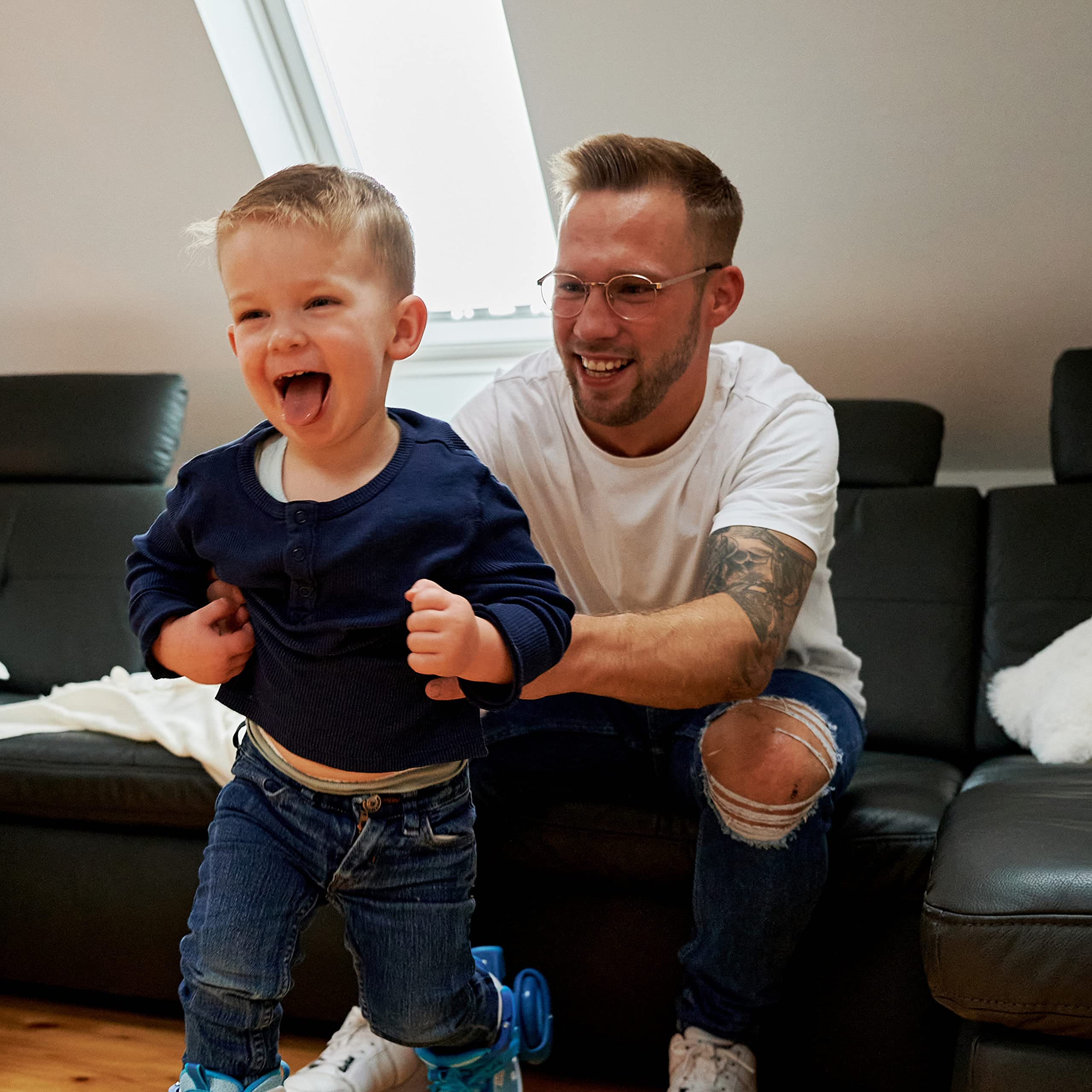 Ein glücklicher Junge in blauer Kleidung läuft, während sein Vater ihn auf einer schwarzen Couch unterstützt.