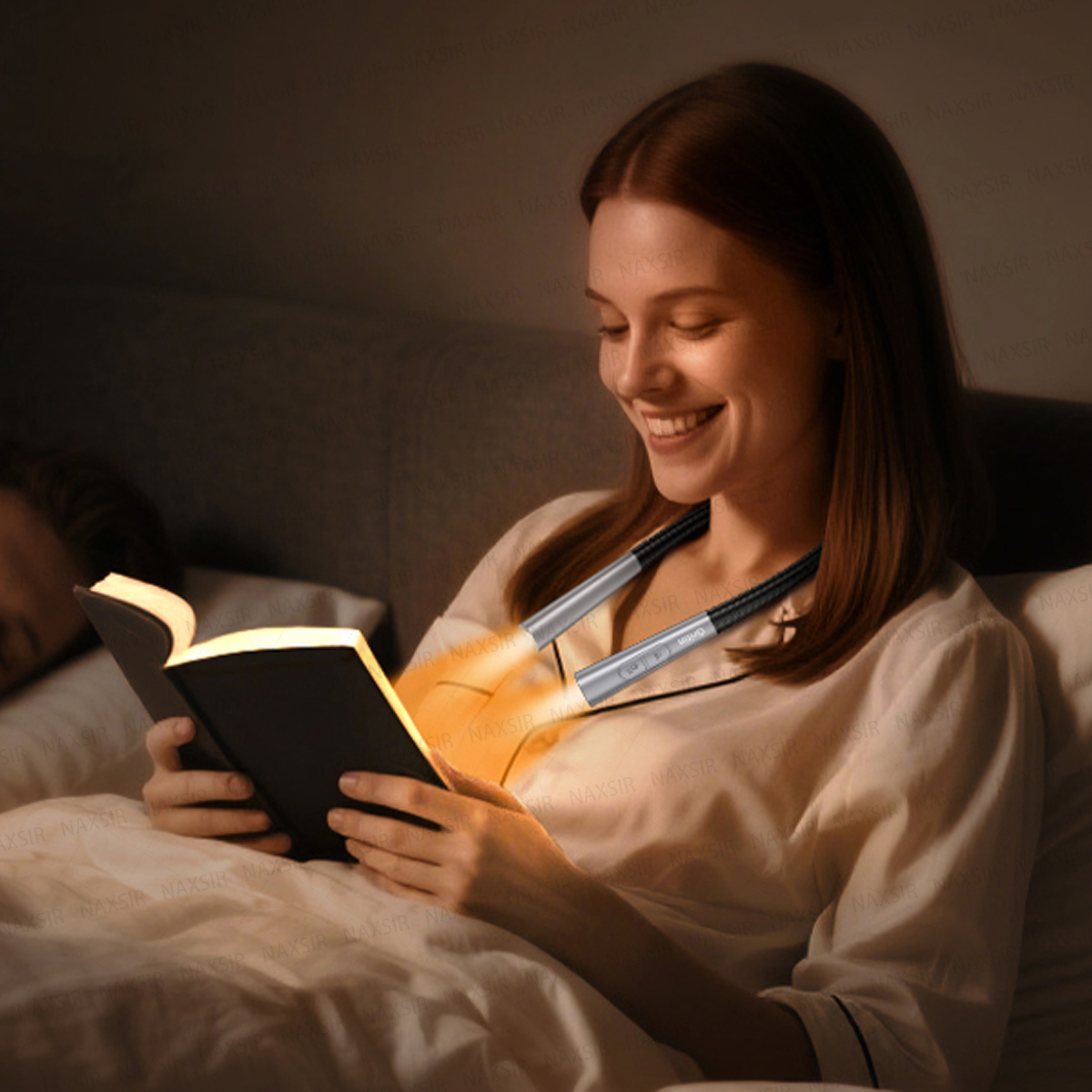 Eine lächelnde Frau im Bett liest ein Buch, beleuchtet von einer Nackenlampe.
