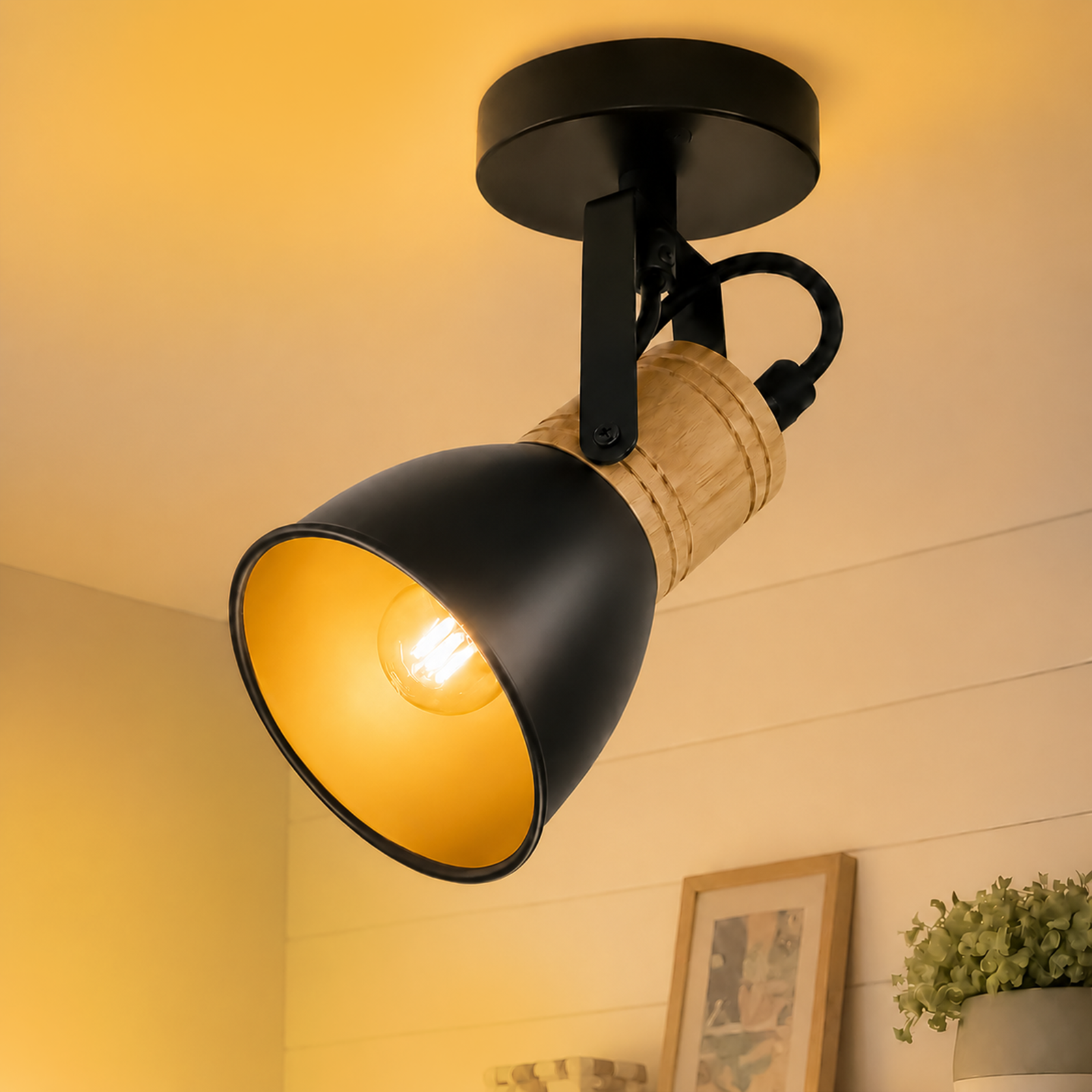 Un foco de techo negro y de madera ilumina una habitación con una bombilla brillante, junto a un cuadro y una planta.