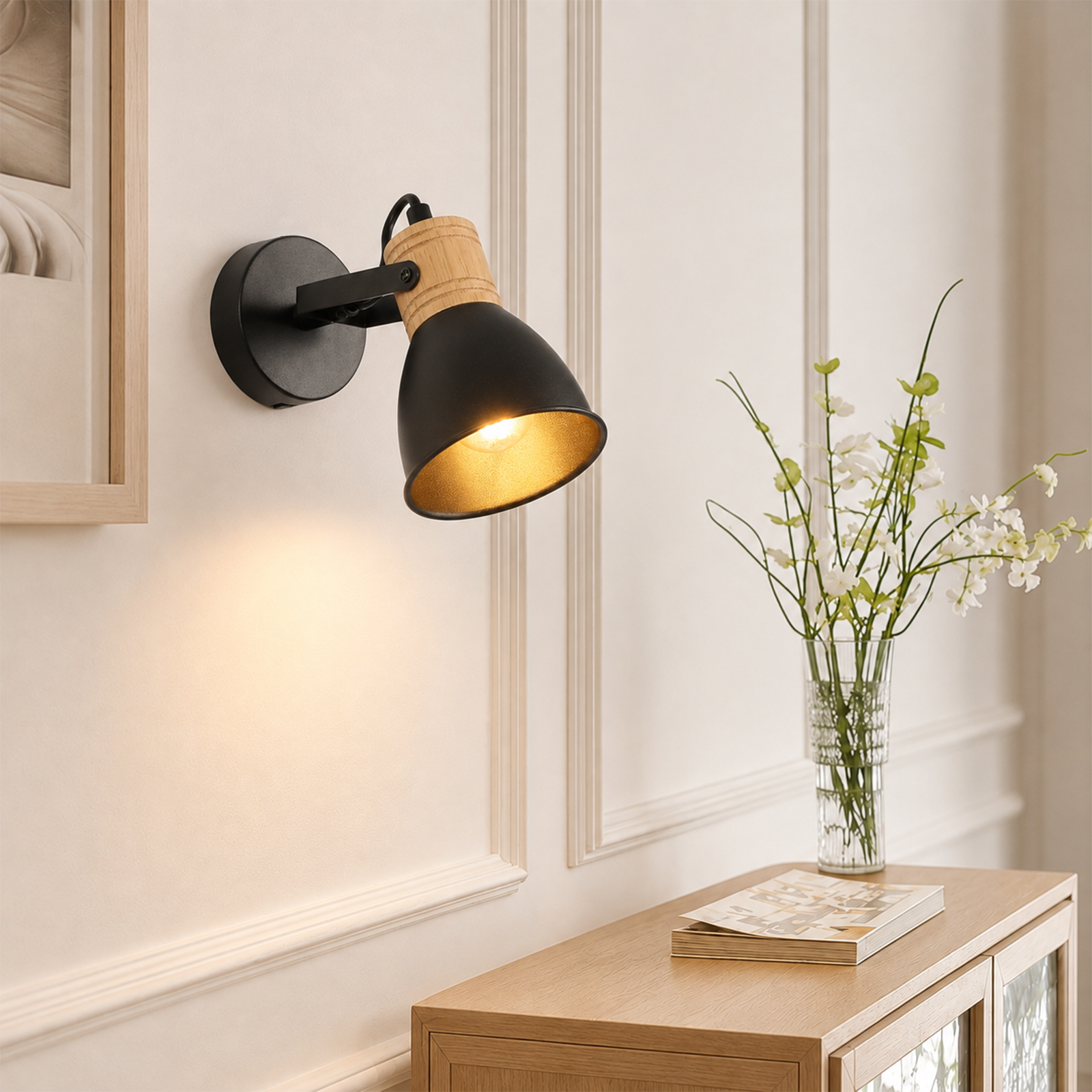 Una lámpara de pared negra y de madera brilla en una pared con paneles, junto a un pequeño armario con flores.