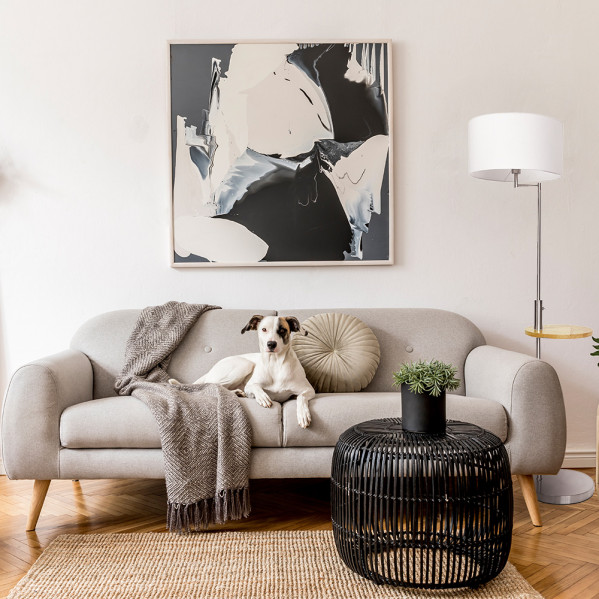Un perro blanco descansa en un sofá gris con manta y cojín redondo, con arte abstracto arriba.