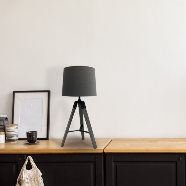 Una lámpara de mesa gris con base de trípode de madera sobre una mesa de madera ante una pared blanca, con un cuadro al lado.
