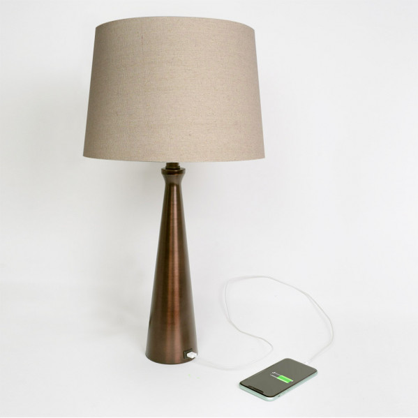 Una lámpara de bronce con pantalla beige sobre mesa blanca, conectada a un teléfono mediante cable.