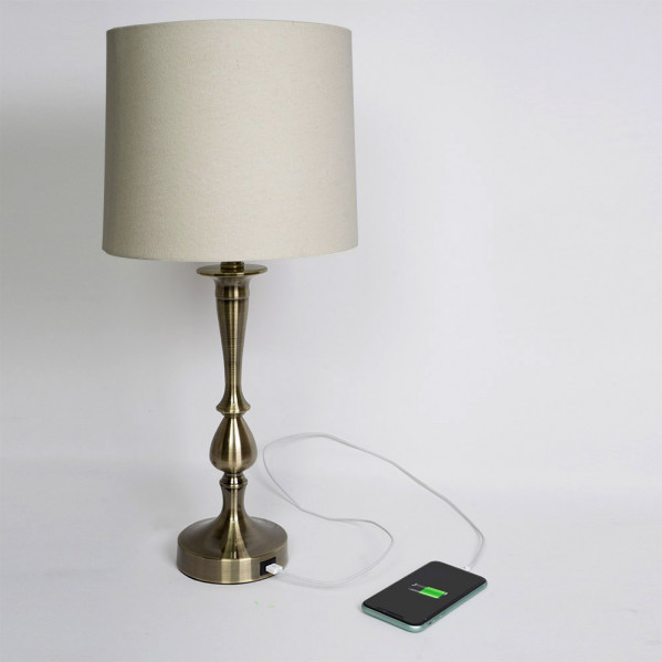 Una lámpara de mesa de latón con pantalla beige carga un teléfono inteligente mediante un cable conectado a su base.