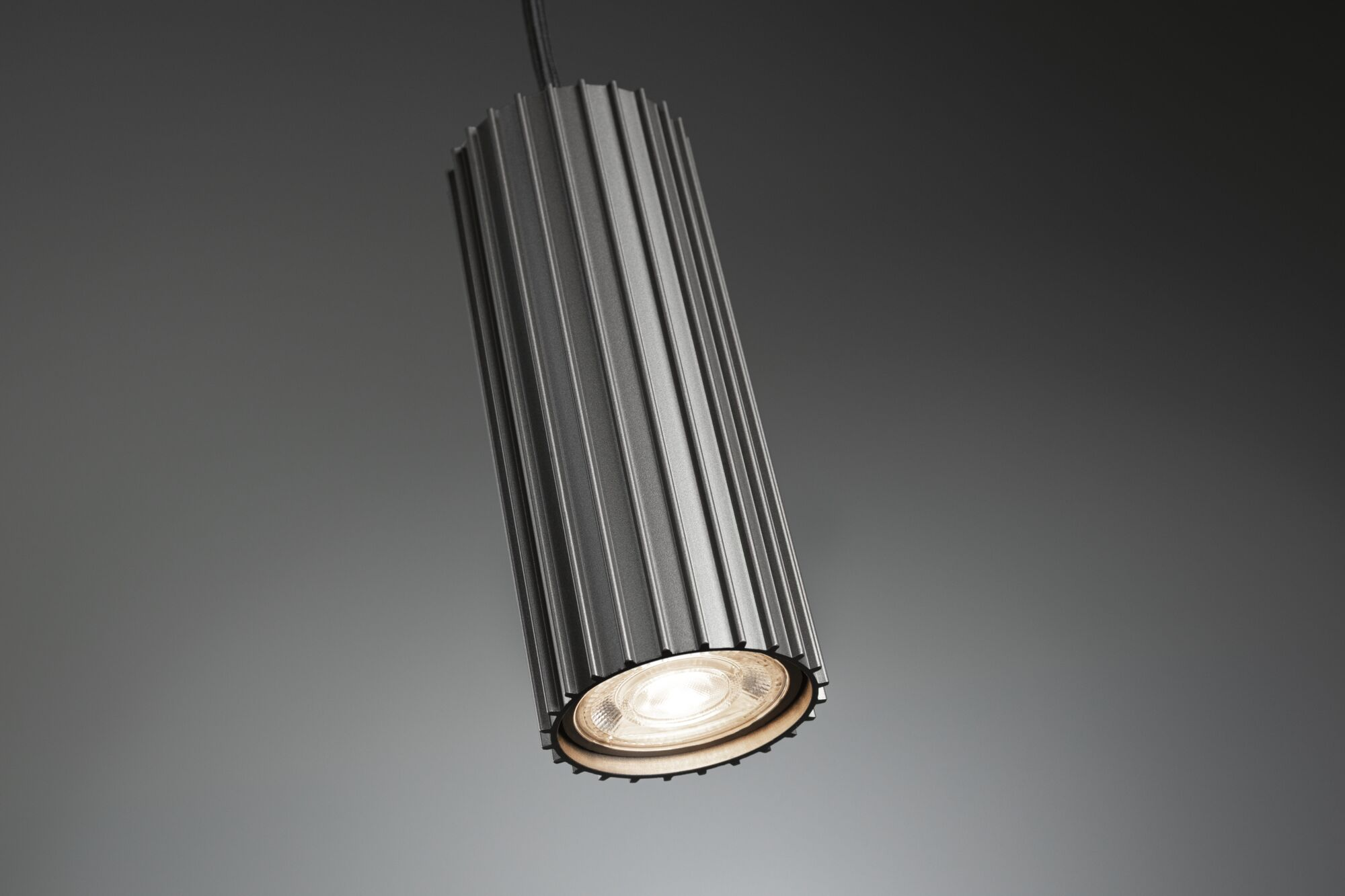 Eine graue, zylindrische Hängeleuchte mit geripptem Design und eingeschaltetem Licht vor grauem Hintergrund.