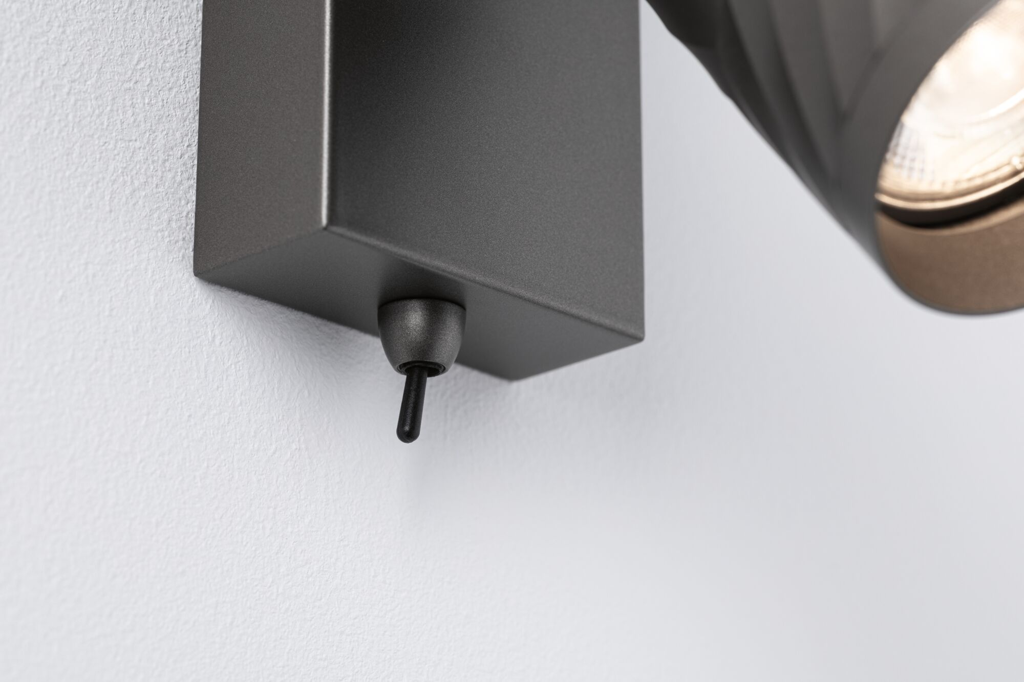Eine schwarze Wandleuchte mit Kippschalter an einer weiß strukturierten Wand.