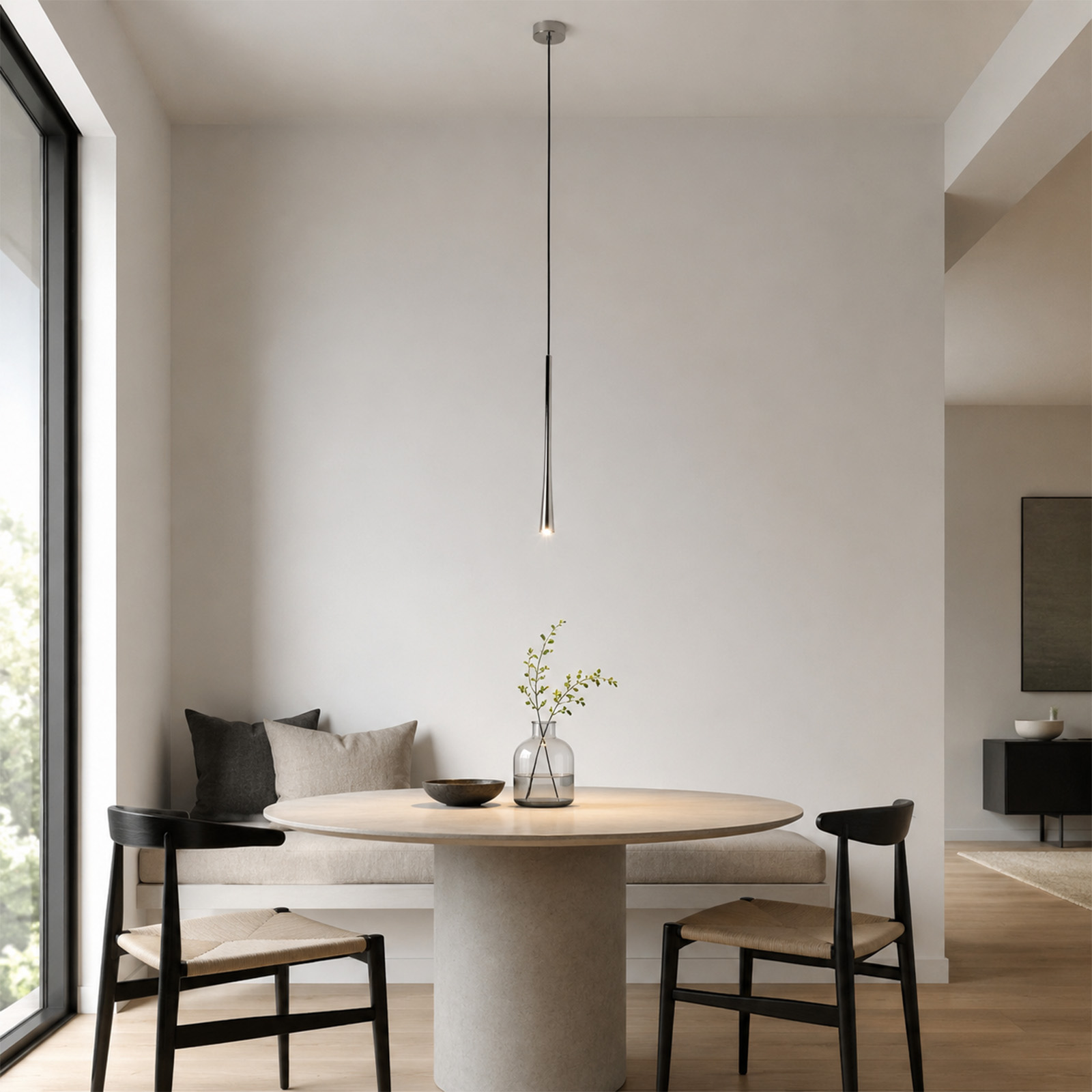 Una mesa de comedor redonda con dos sillas negras y un banco empotrado, iluminada por una lámpara colgante delgada.