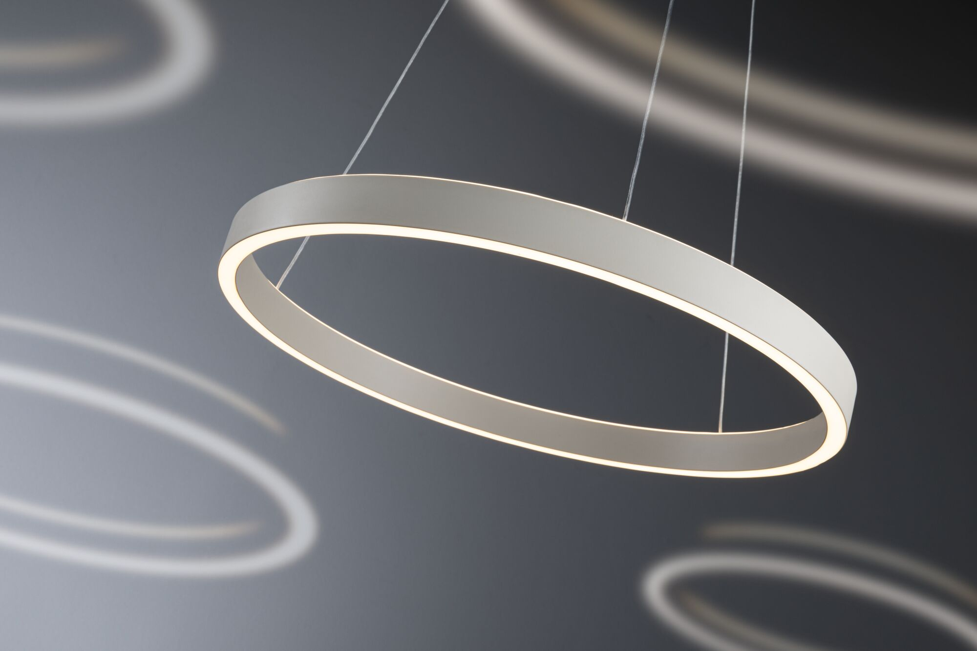 Eine moderne weiße Hängeleuchte in Ringform leuchtet vor einem grauen Hintergrund mit kreisförmigen Lichtreflexen.