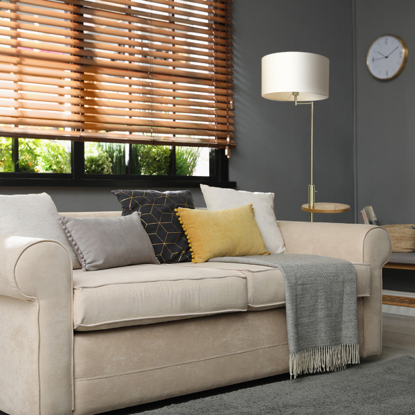 Sofá beige con cojines y manta, lámpara de pie, persianas de madera y reloj de pared.