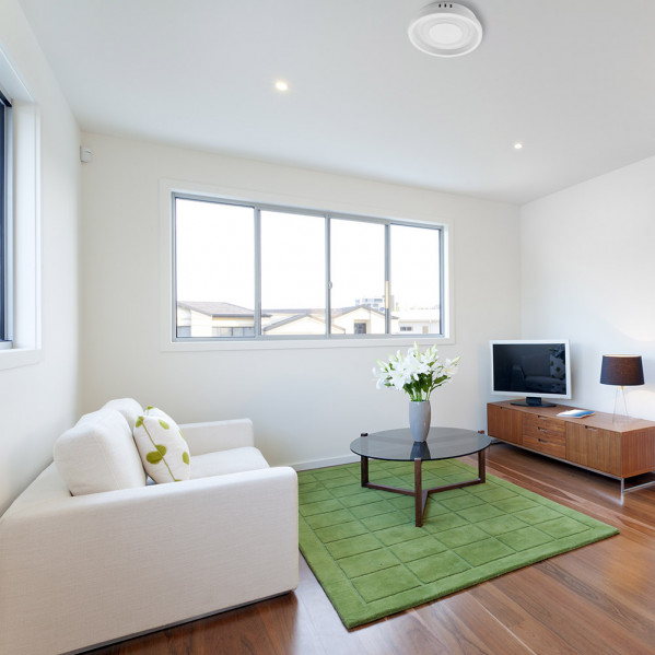 Una sala luminosa tiene sofá blanco, alfombra verde, mesa de centro redonda con flores, televisor y mueble.