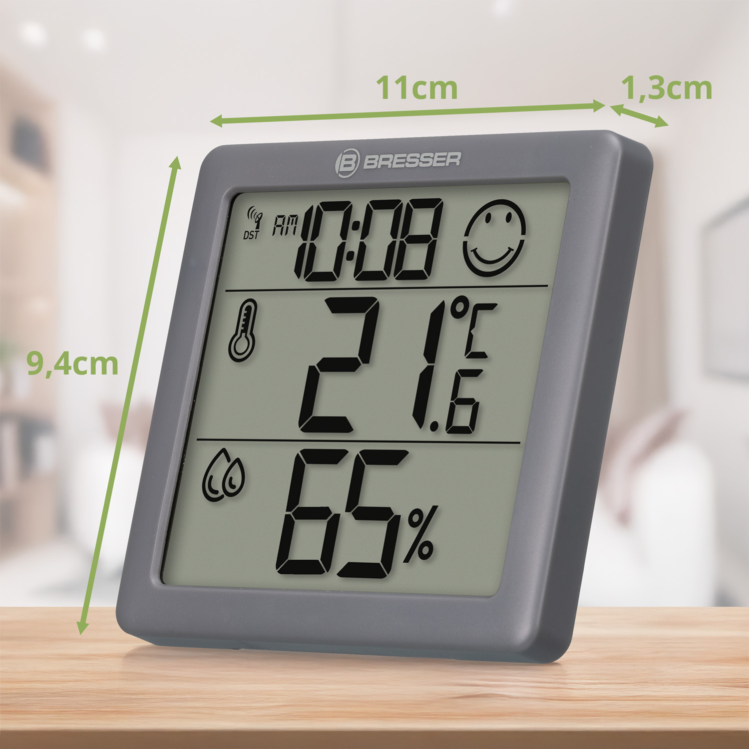 Reloj meteorológico digital gris que muestra hora, temperatura y humedad con flechas de dimensión en una mesa.