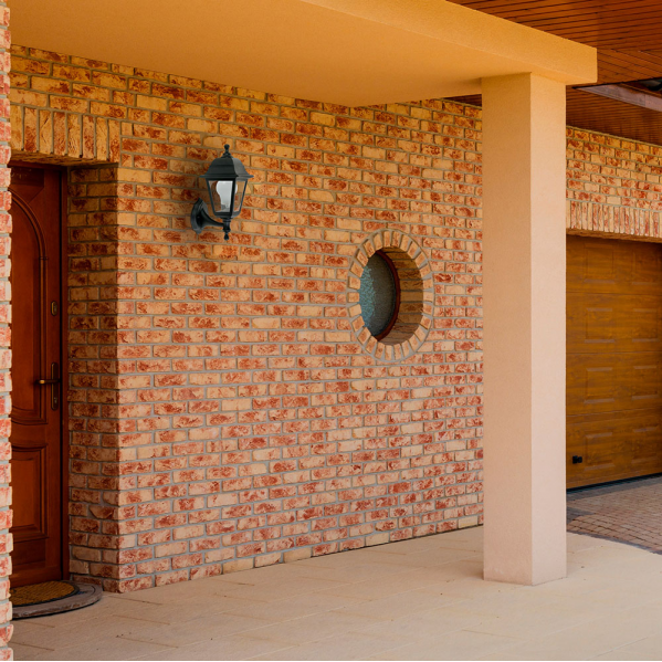 Una lámpara de exterior negra en una pared de ladrillo rojo junto a una puerta de madera y una ventana redonda.