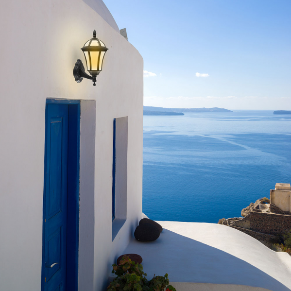 Un farol de pared iluminado en un edificio blanco con puerta azul frente al mar.