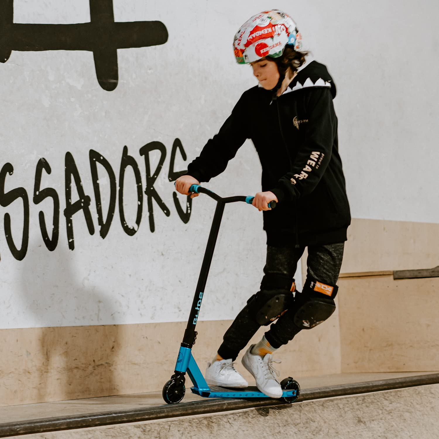 Ein Kind mit schwarzem Hoodie, Helm und Knieschonern fährt auf einer Rampe mit Graffiti-Hintergrund Stunt-Scooter.