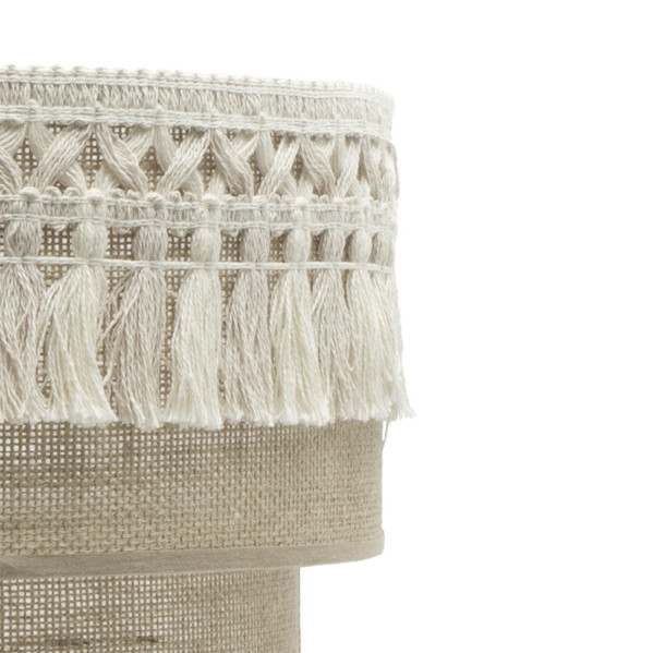 Primer plano de una pantalla de lámpara de tela beige decorada con cenefa de macramé blanca y borlas sobre fondo blanco.