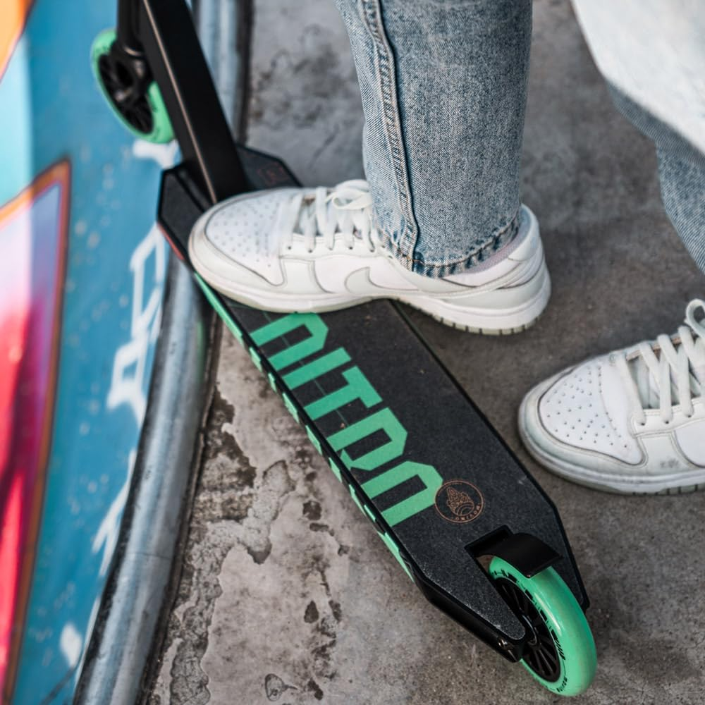 Person in Jeans und weißen Sneakers steht auf einem schwarzen Stunt-Scooter mit grünen Akzenten.