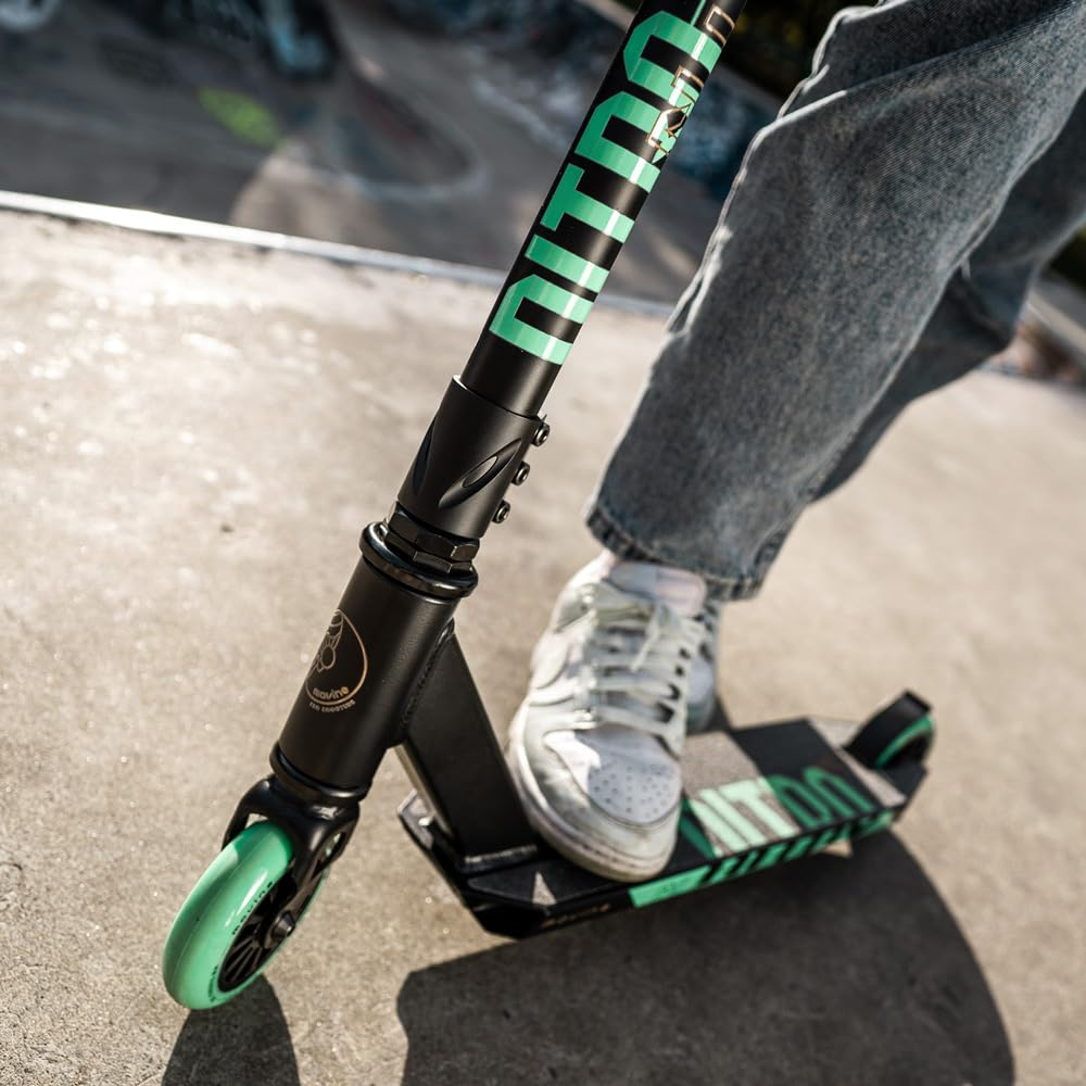 Person in Jeans und weißen Sneakers steht auf einem schwarz-türkisen Stunt-Scooter auf Beton.